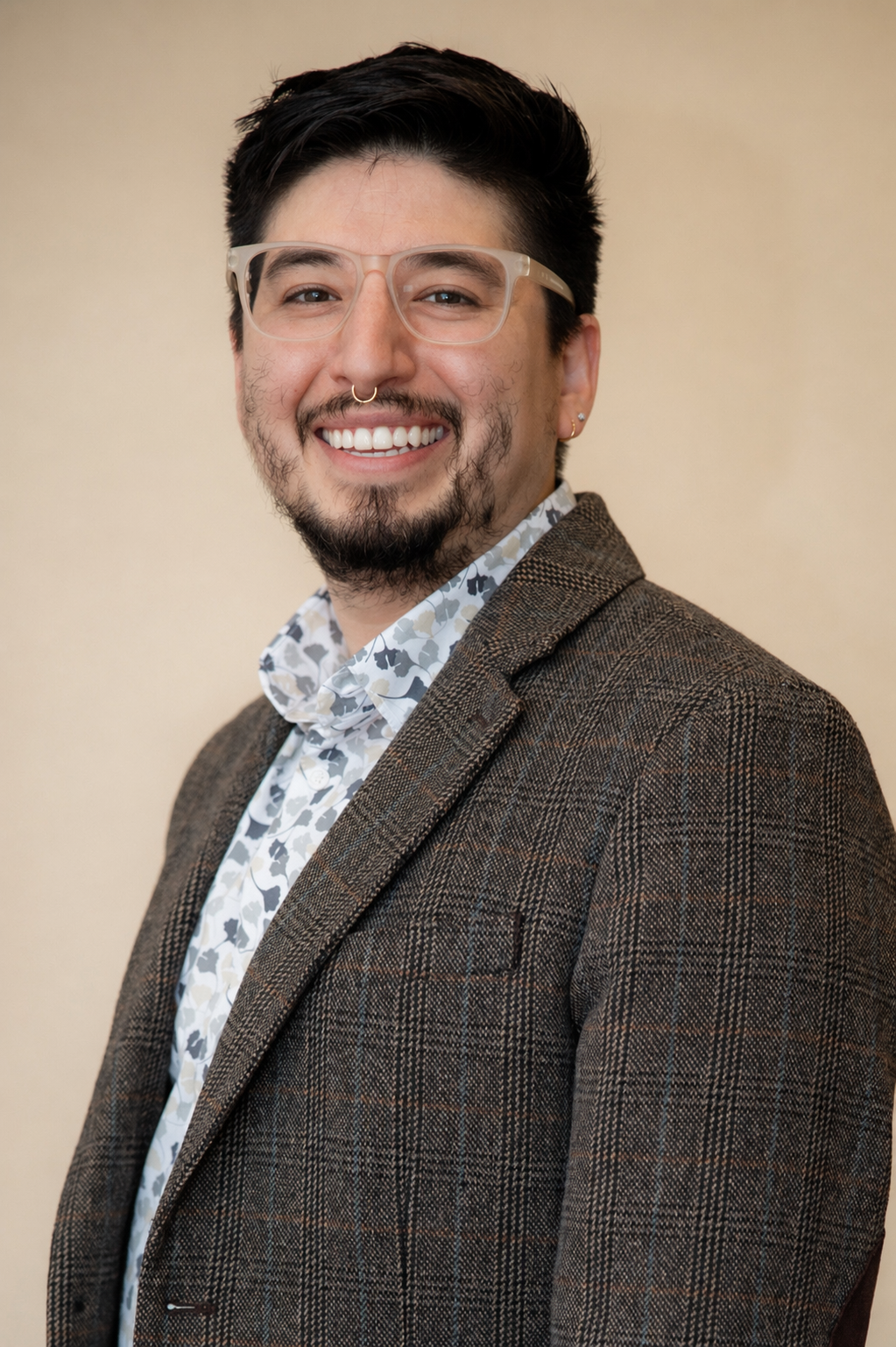 A man with glasses, a septum piercing, and earrings, smiling and wearing a checkered blazer over a patterned shirt, with a plain, light-colored background.