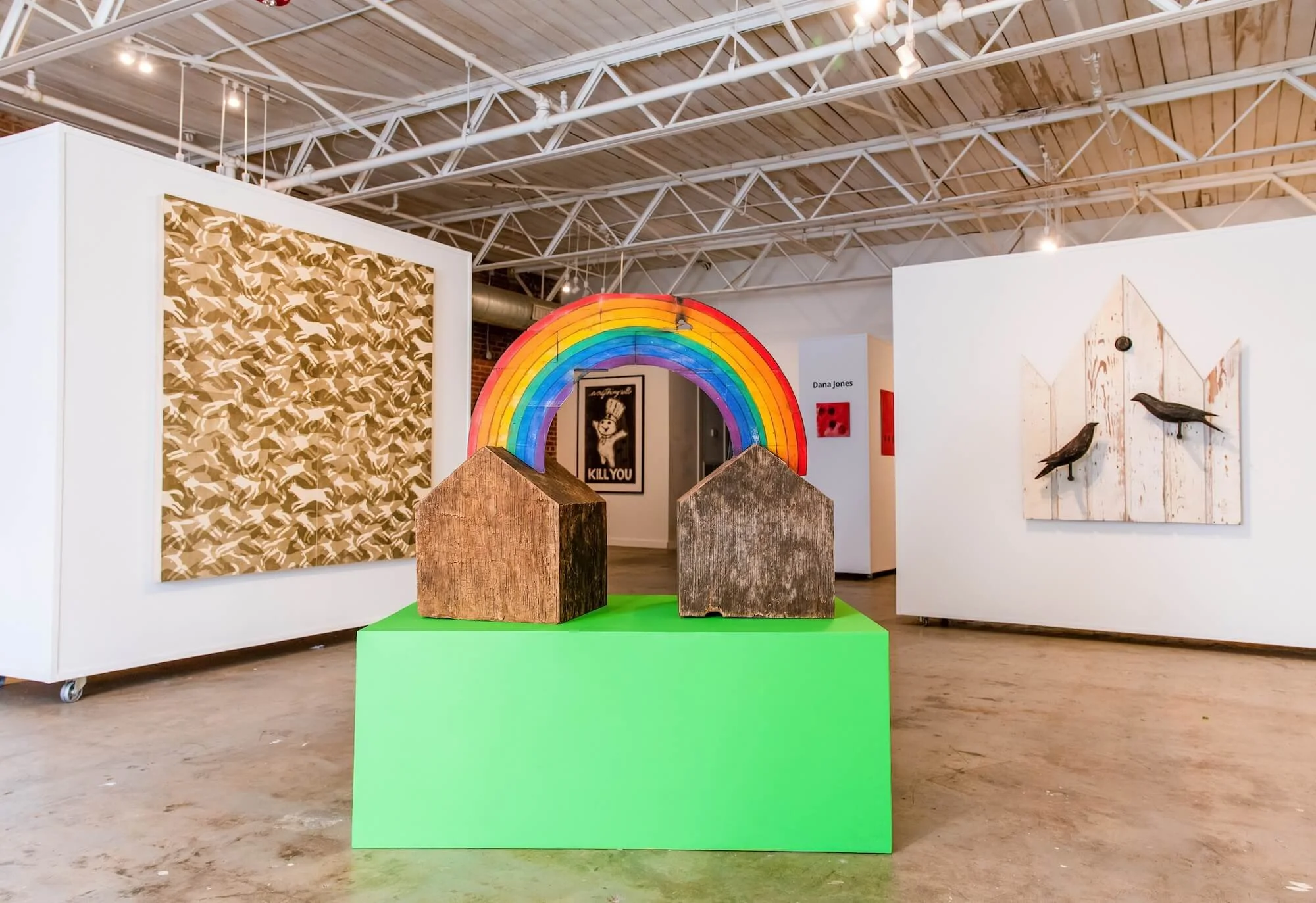 Art gallery showcasing various artworks, including a colorful rainbow sculpture with two wooden house structures underneath, a patterned painting of running animals on the left, and a wood art piece with two black birds on the right wall.