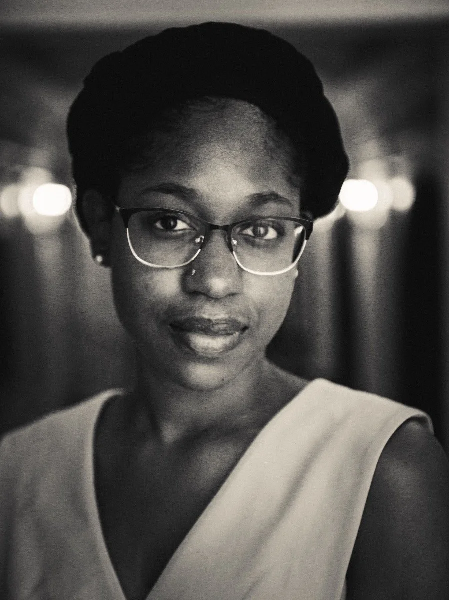 A close-up black-and-white portrait of a woman with glasses, short hair styled in a twist, wearing a sleeveless top, looking directly at the camera.