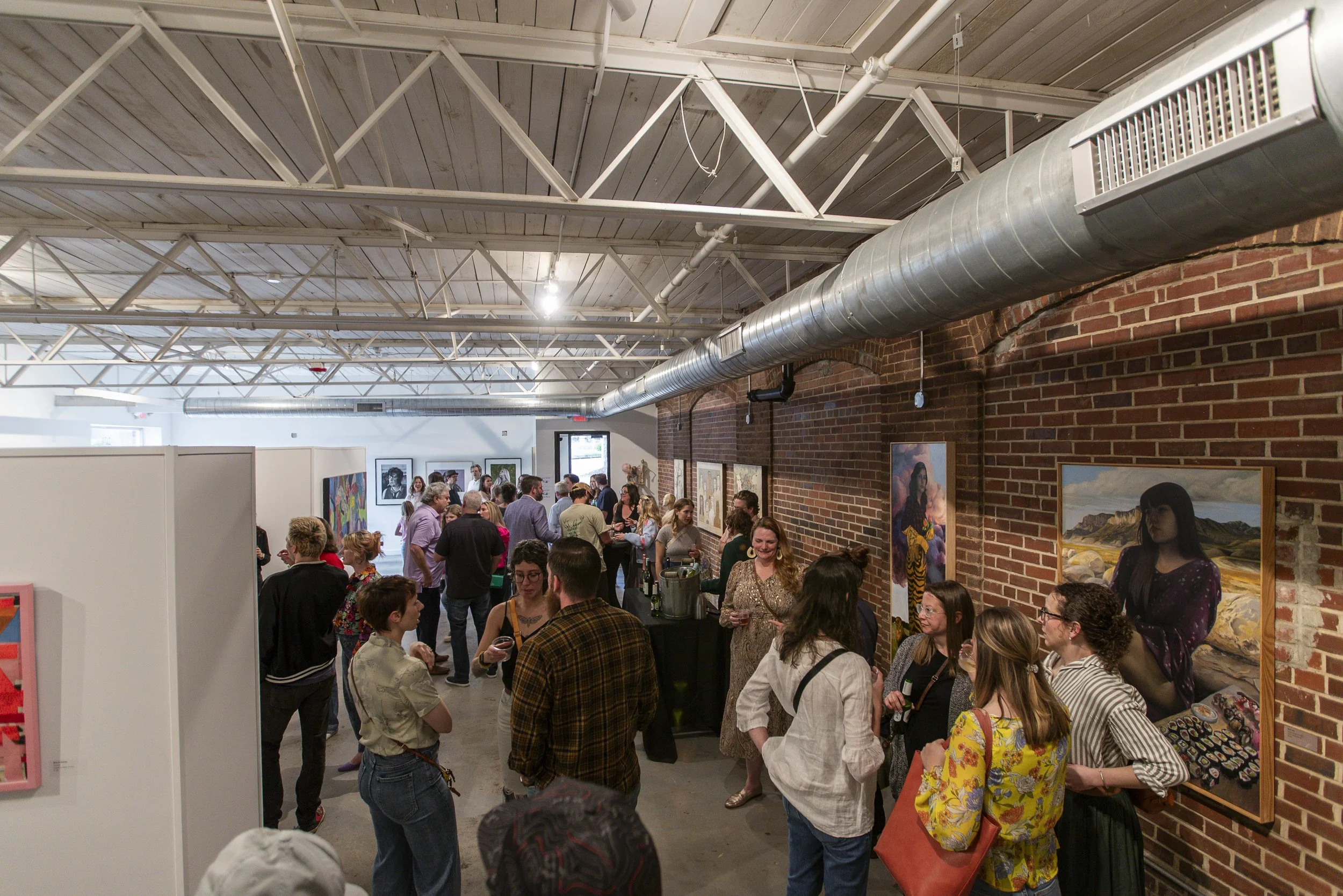 People at an art gallery viewing various paintings on brick walls and white partitions during an event.
