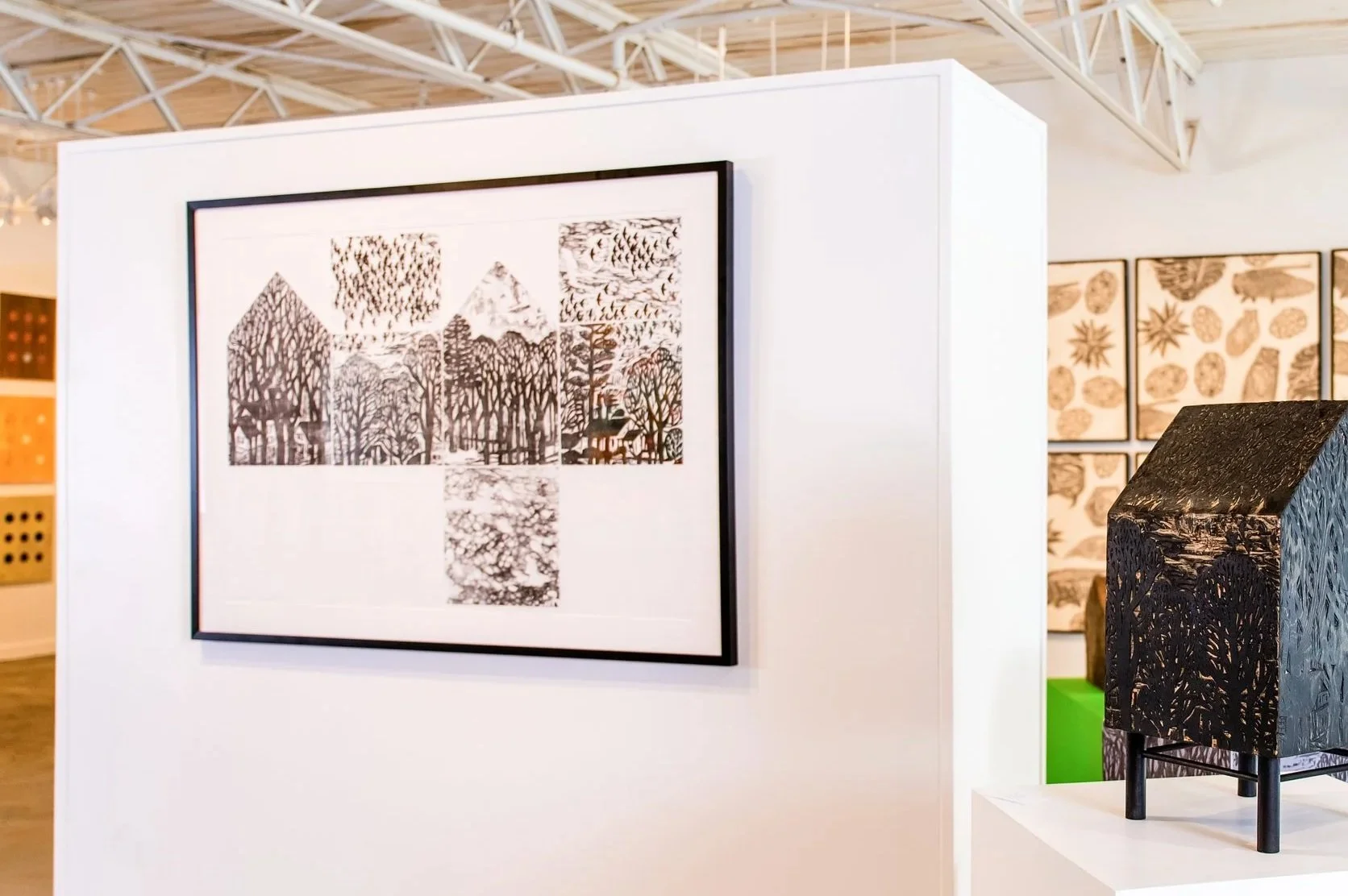 Art gallery display with a framed artwork featuring a black and white forest scene and abstract shapes. A black textured sculpture on a white pedestal is nearby in the exhibit space.