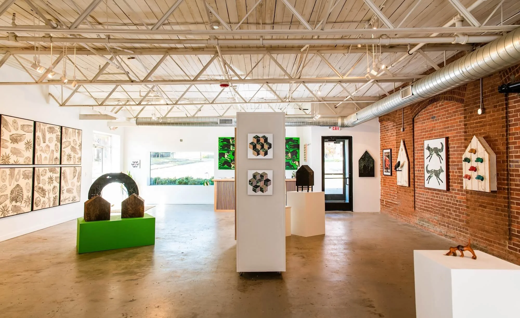 Interior of an art gallery with various artworks on white and brick walls, sculptures, and bright natural lighting.