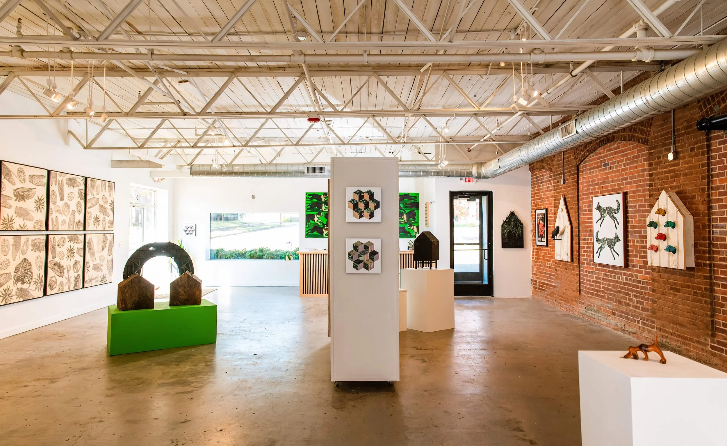Art gallery interior with paintings, sculptures, and mixed media artworks on brick and white walls, with large windows providing natural light, and a polished concrete floor.