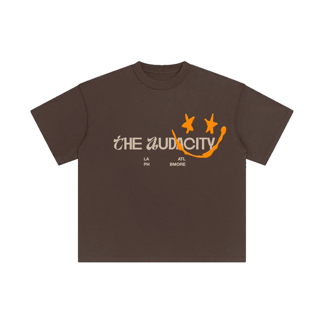 Brown T-shirt with the text 'The Mid City' and a stylized smiley face graphic in orange, with stars for eyes and a curved line for a mouth. The T-shirt has small text below the main text indicating locations: 'LA', 'PH', 'ATL', 'B MORE'.