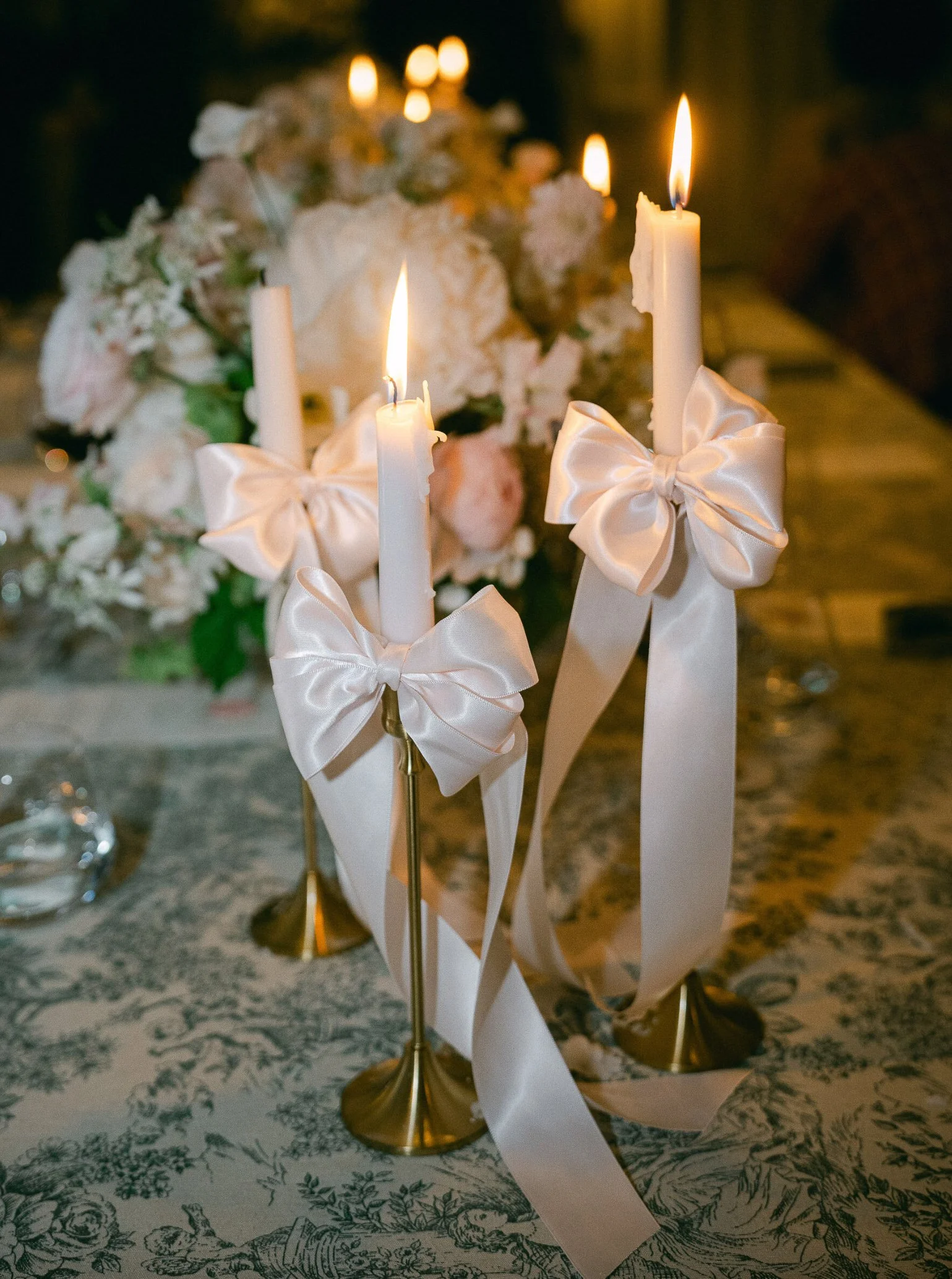 A romantic candlelit table centerpiece with three lit white candles decorated with large white satin bows, with a background of flowers and candles, on a floral tablecloth.