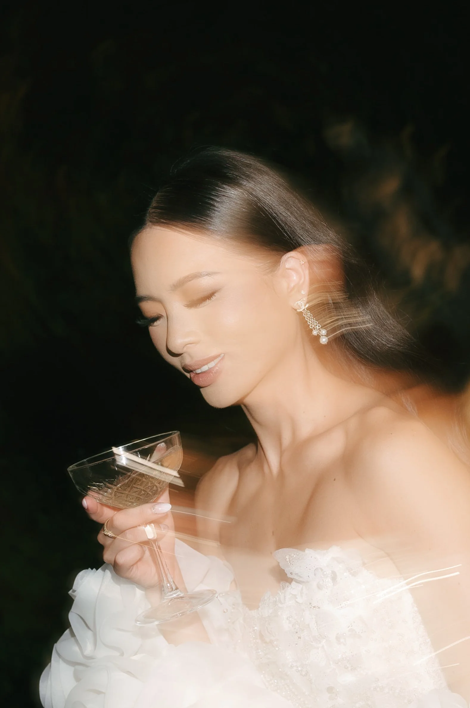 A woman in a white dress with lace details holding a champagne glass, elegant earrings, and a ring, with dark background.