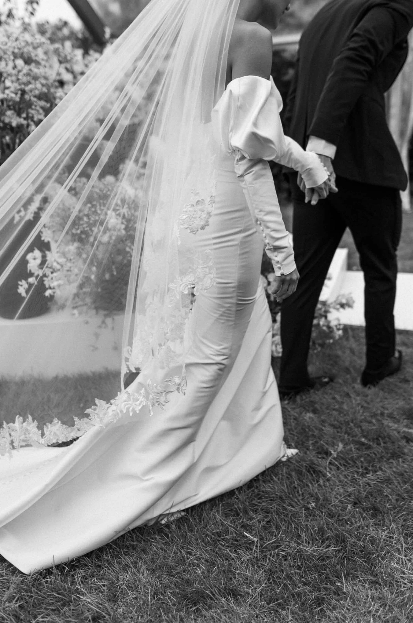 Black and white photo of a bride and groom holding hands during a wedding ceremony outdoors. The bride is wearing a long satin gown with lace details and a veil, while the groom is dressed in a dark suit. They are standing on grass with flowers in the background.