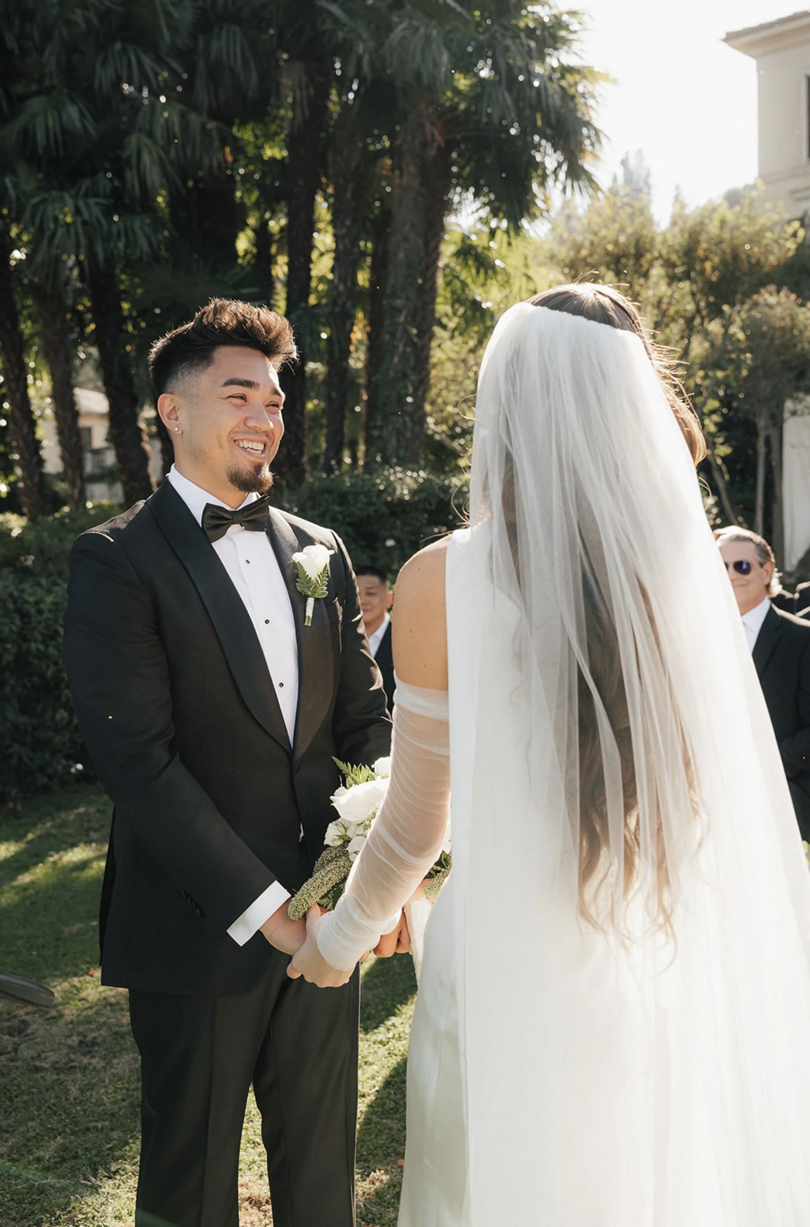 A groom in a black tuxedo and bow tie smiling at a bride in a white wedding dress and veil during an outdoor wedding ceremony.