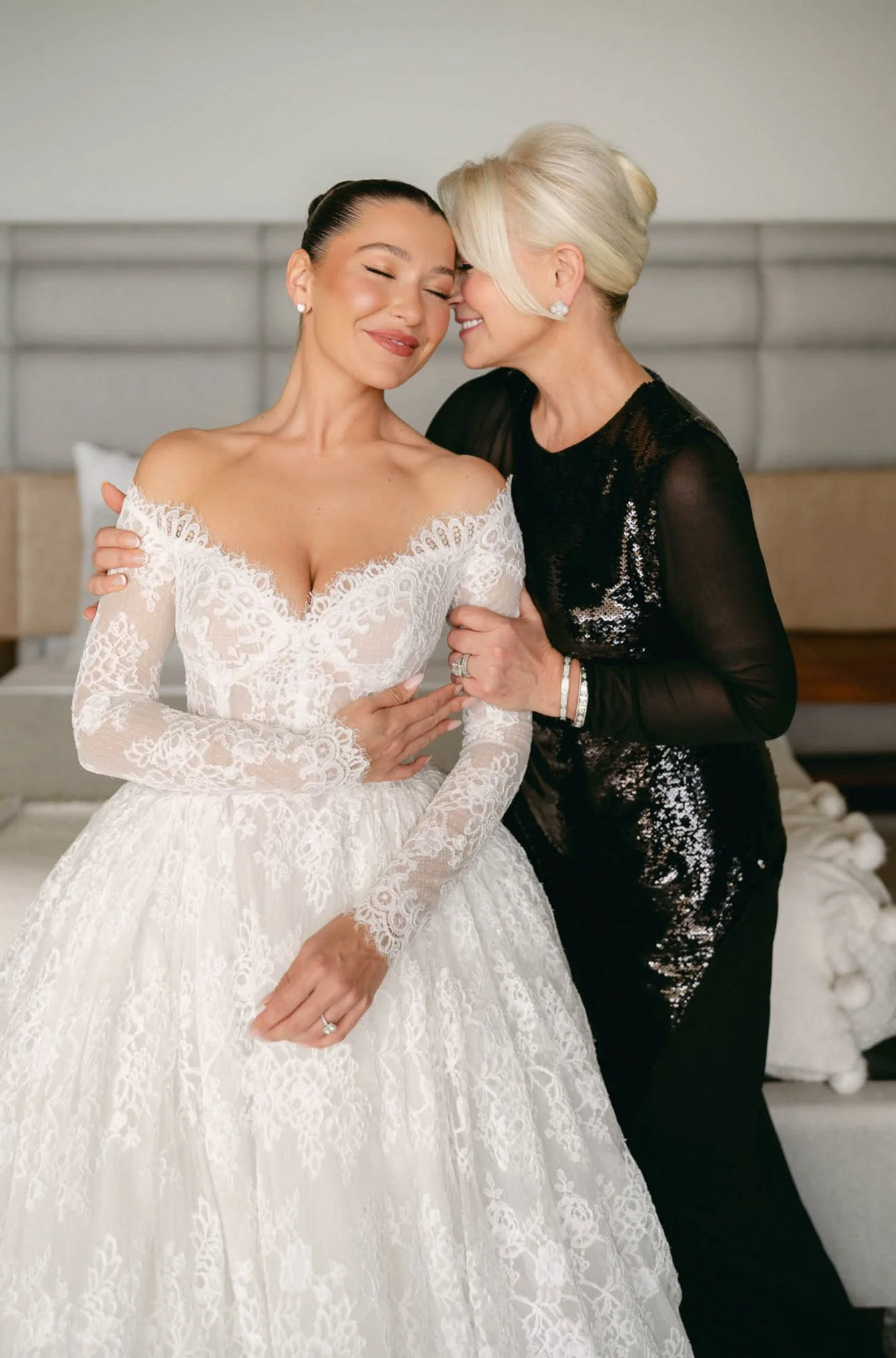 A bride in a white lace wedding dress with off-the-shoulder sleeves and long sleeves, and an older woman in a black sequined dress sharing a joyful moment with their foreheads touching.
