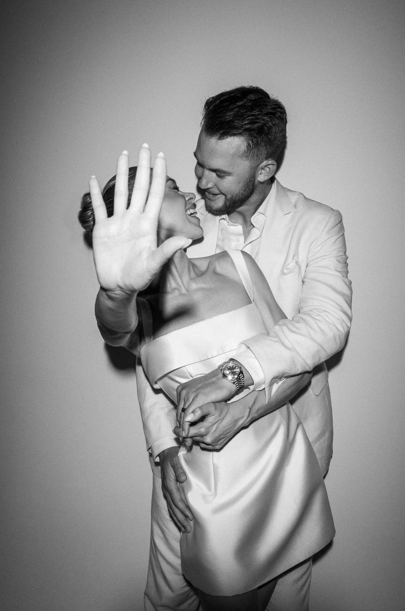 A black-and-white photo of a man and woman smiling and embracing. The woman has her hand raised, partly covering her face, and the man is holding her around the waist. They appear happy and close.