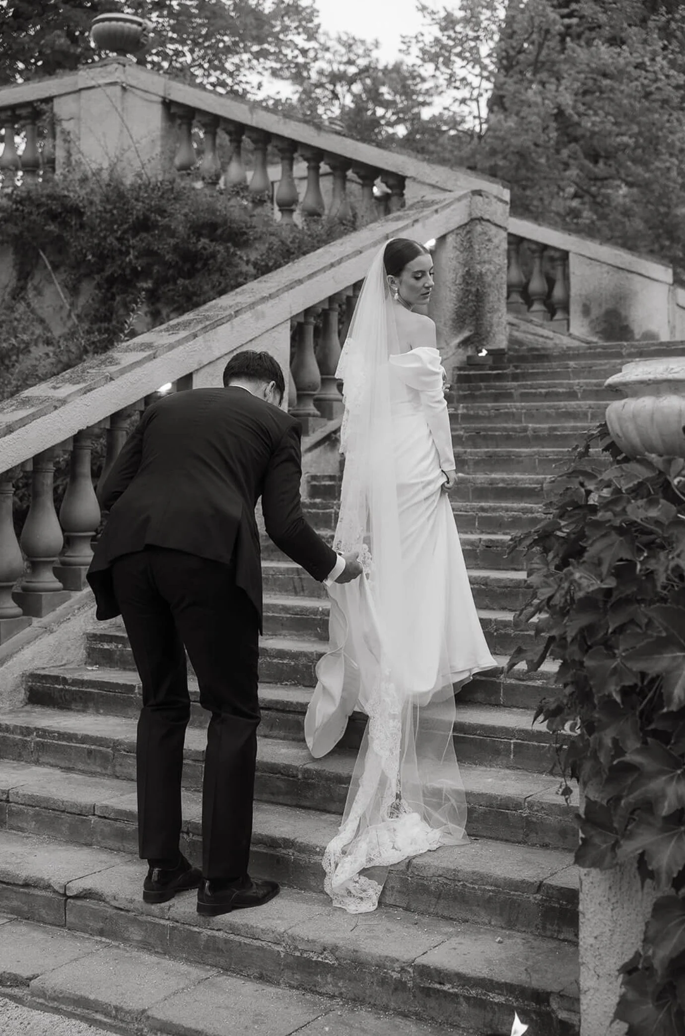 A bride in a white wedding gown and veil stands on outdoor stone stairs, while a man in a dark suit adjusts her dress.
