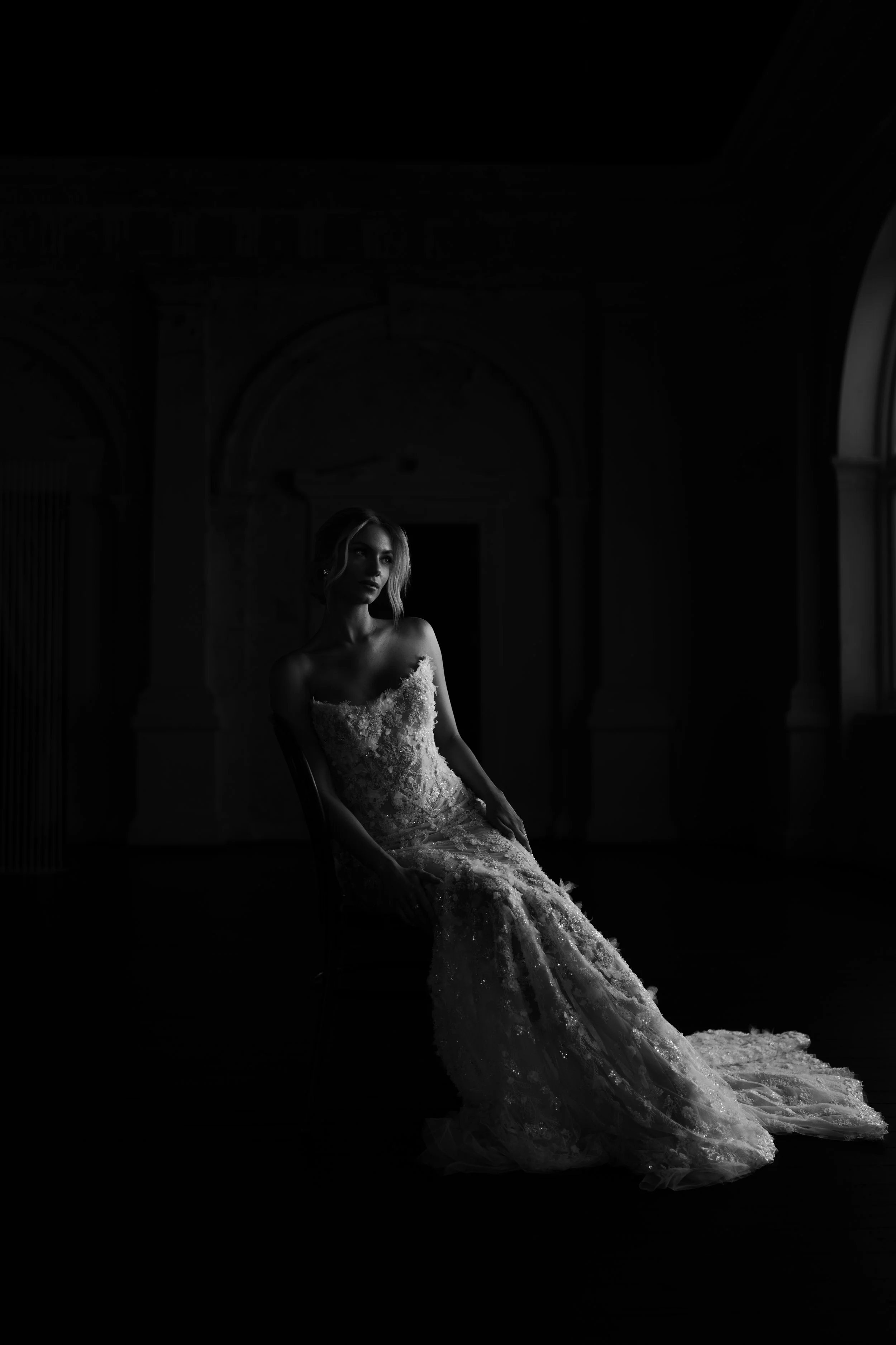 A woman in a wedding dress sitting in a dimly lit room, with light coming from a window on the right.
