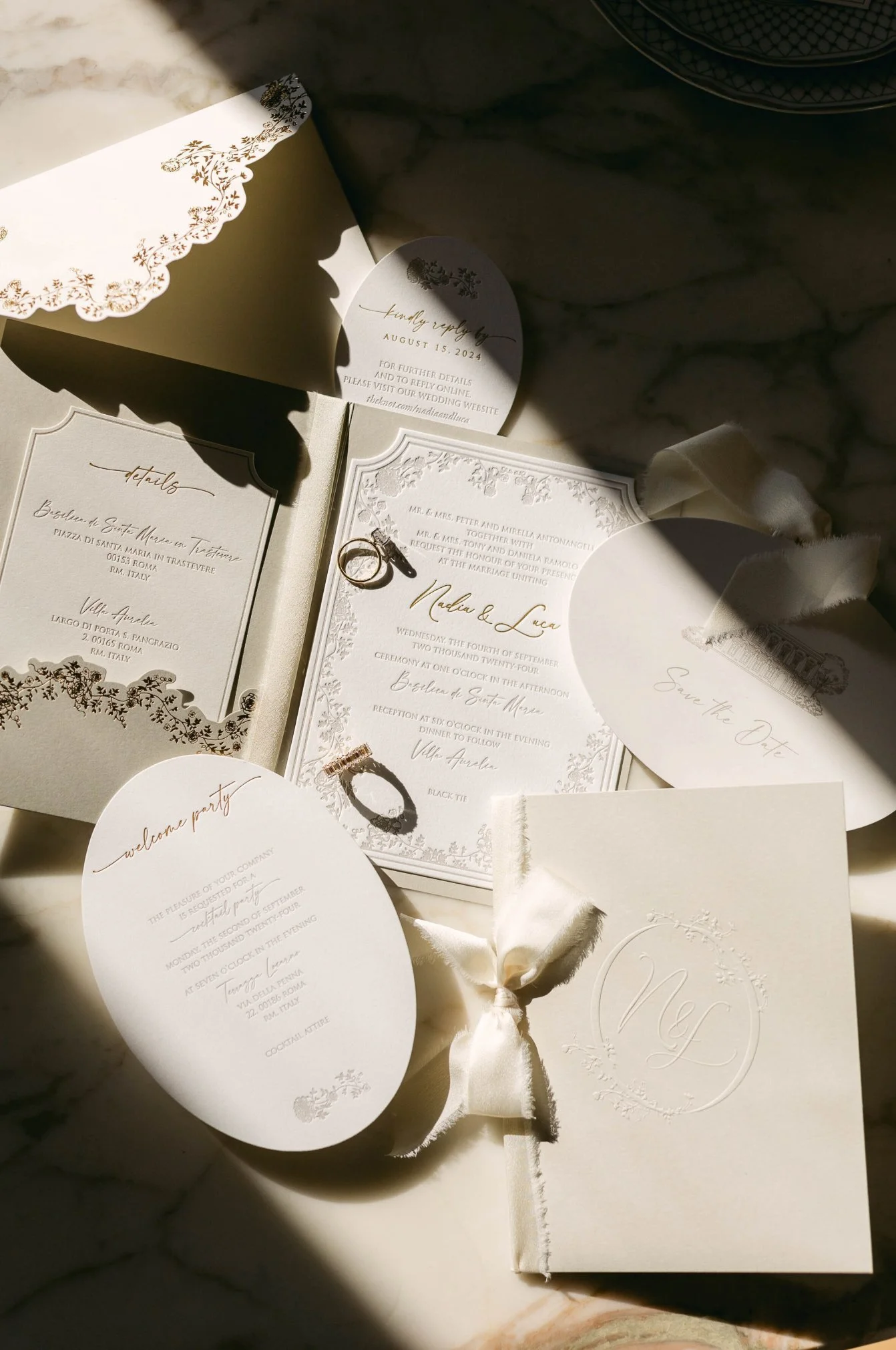 Wedding invitation suite with rings, save the date card, and RSVP card on a marble surface, illuminated by sunlight.