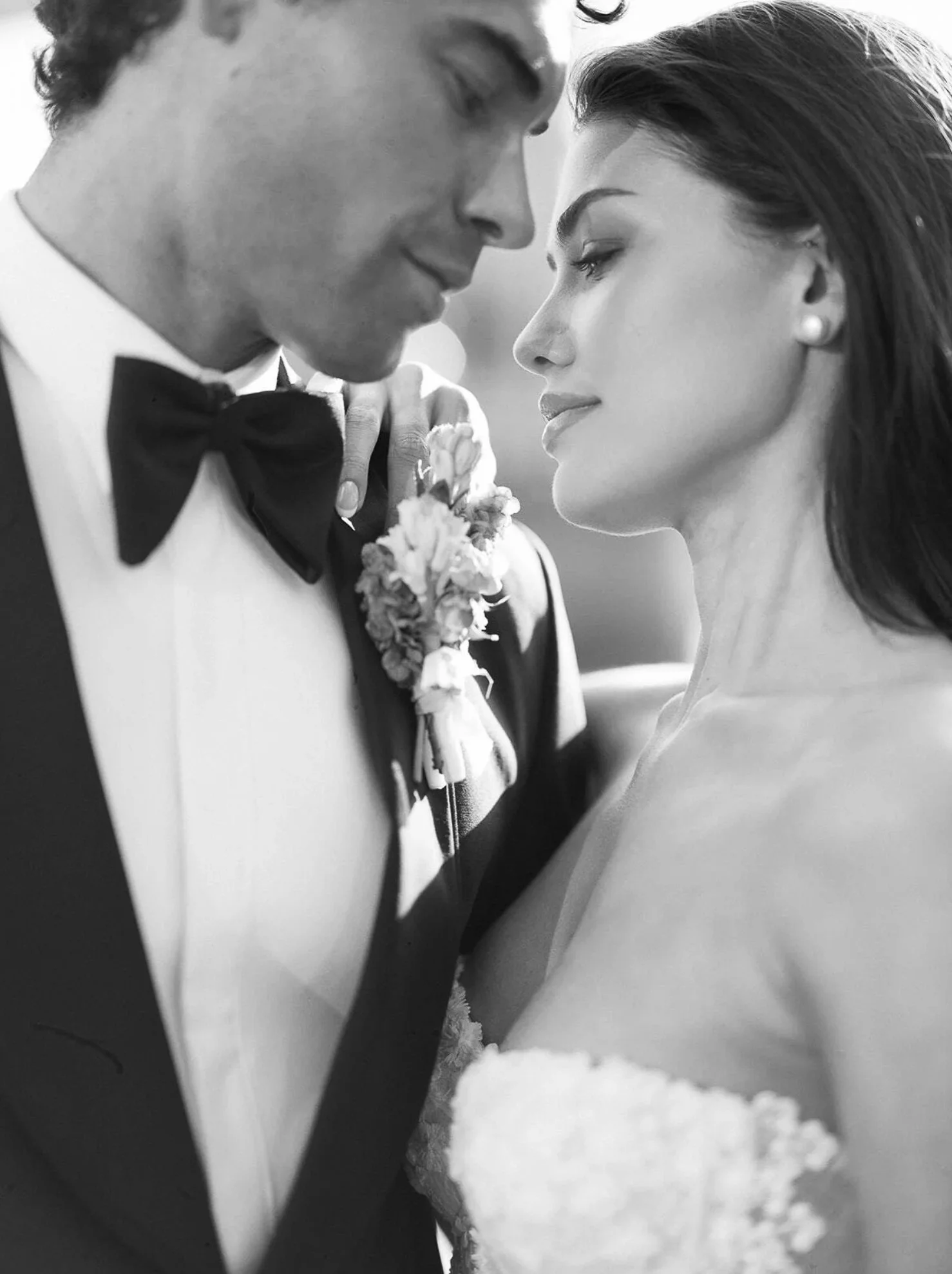 A black and white photo of a bride and groom close together, eyes closed, sharing an intimate moment. The groom is wearing a tuxedo with a bow tie and boutonniere, and the bride is in a strapless wedding dress with pearl earrings.