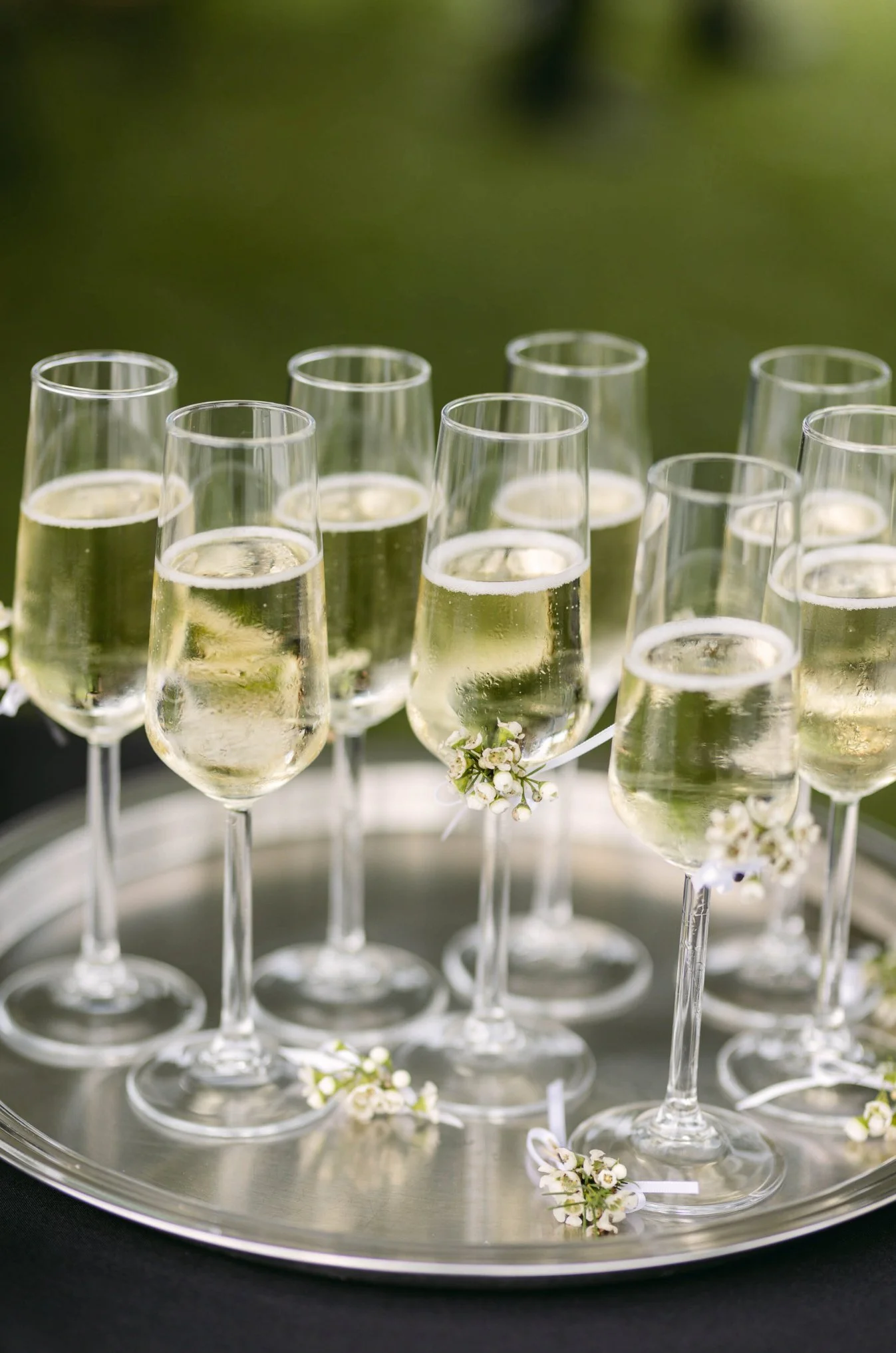 Multiple glasses of champagne on a silver tray, decorated with small white flowers, outdoors.