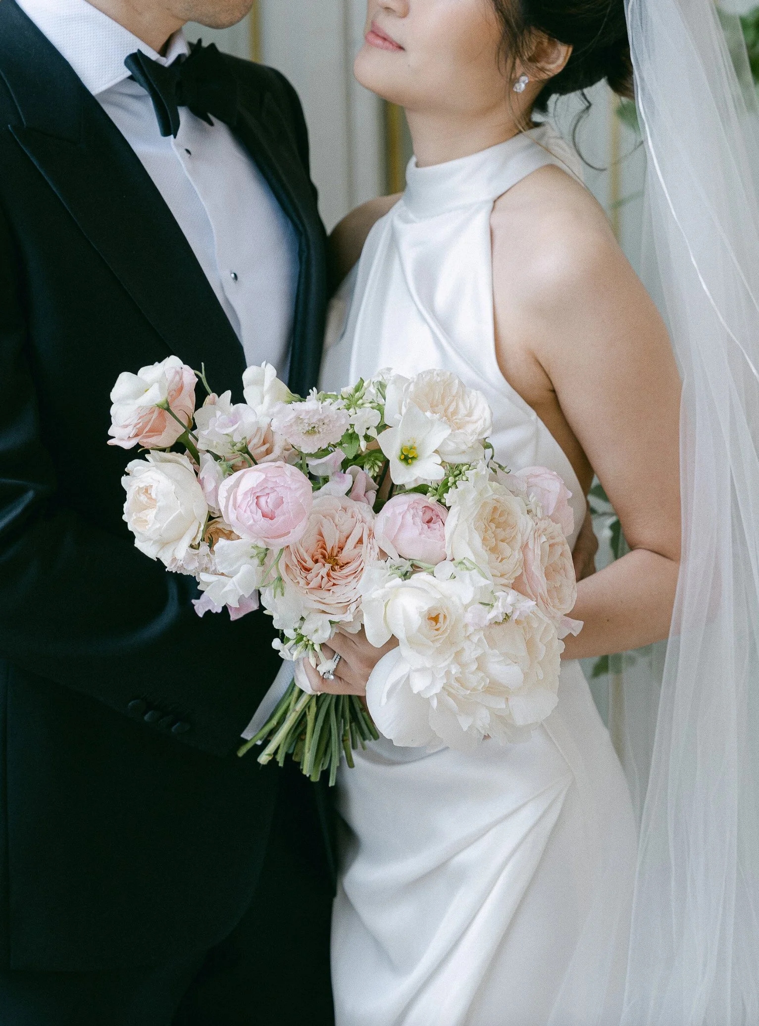 A bride holding a bouquet of white and blush pink flowers. She is wearing a white wedding dress with a high neckline and has a sheer veil. The groom, dressed in a black tuxedo with a bow tie, stands close to her.