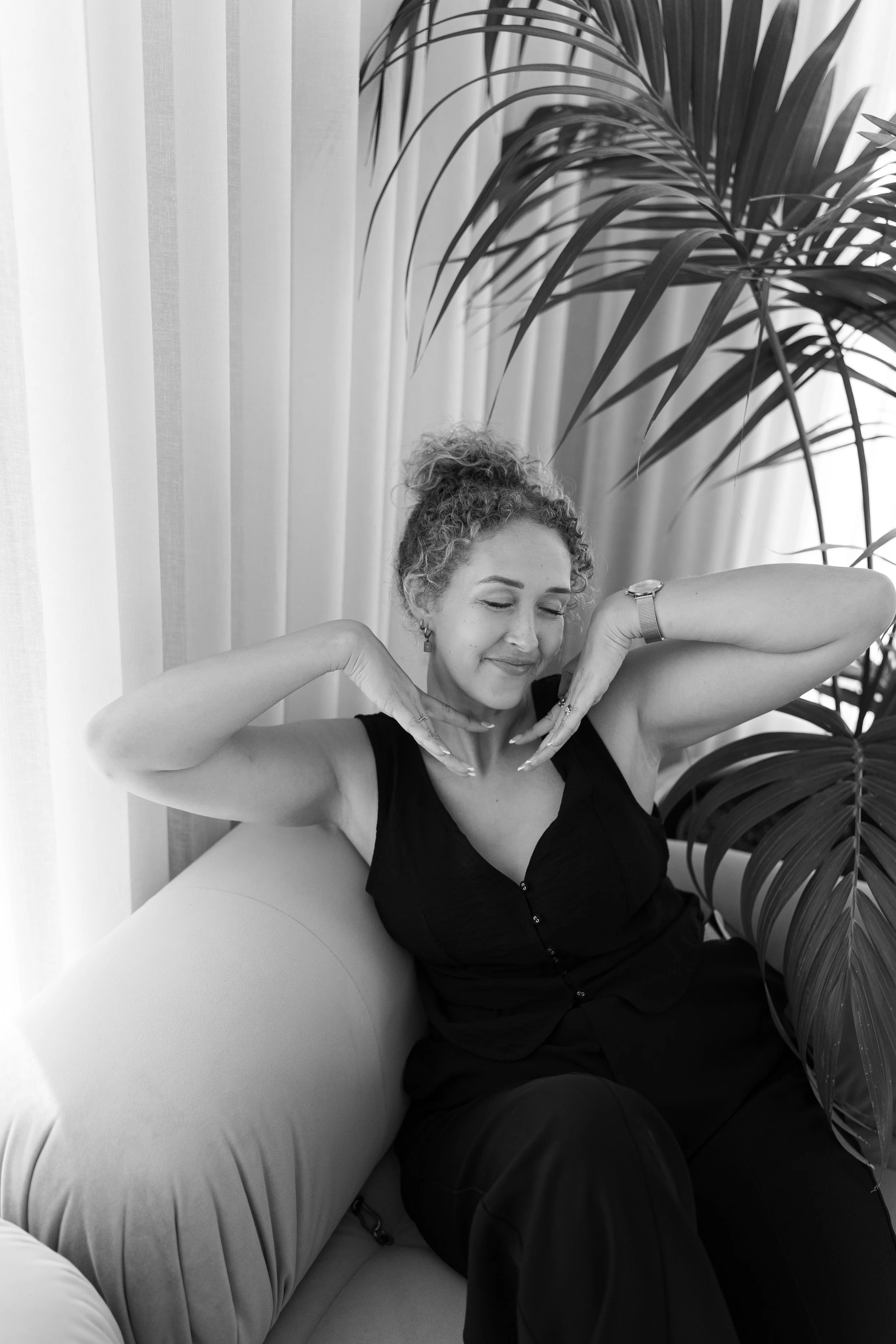 A woman with curly hair sitting on a sofa with her eyes closed. She is wearing a sleeveless black top and has her hands near her face in a relaxed pose. There are large leafy plants and curtains in the background.