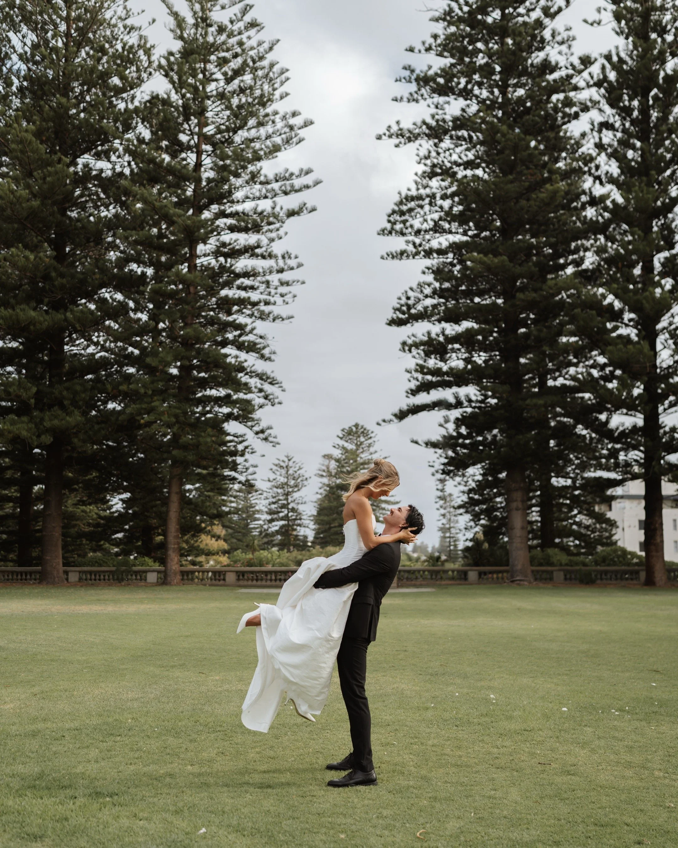 A man in a black suit lifting a woman in a white wedding dress in an outdoor park with tall evergreen trees and a cloudy sky.