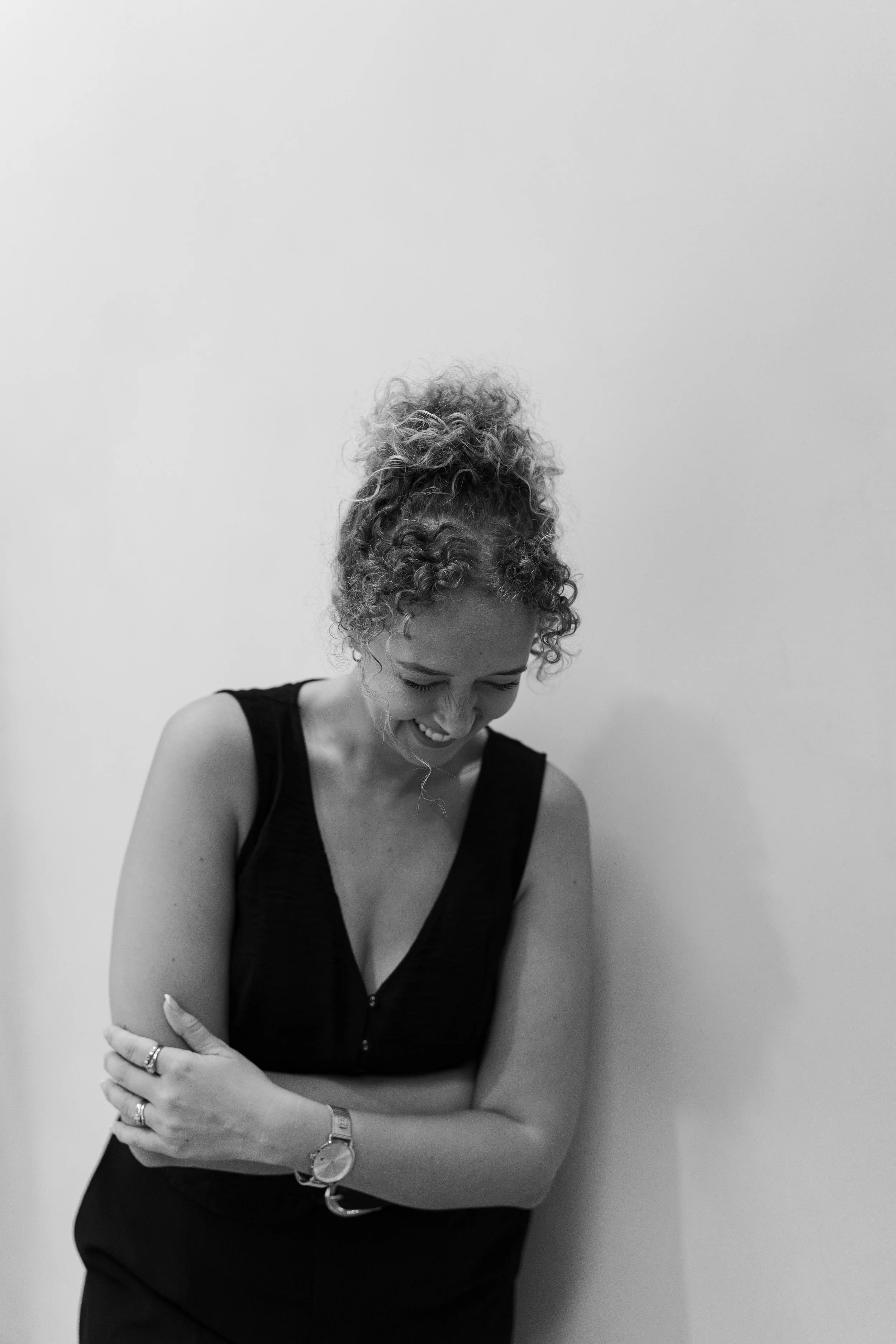 Black and white portrait of a woman with curly hair smiling and looking down, arms crossed, against a plain wall.