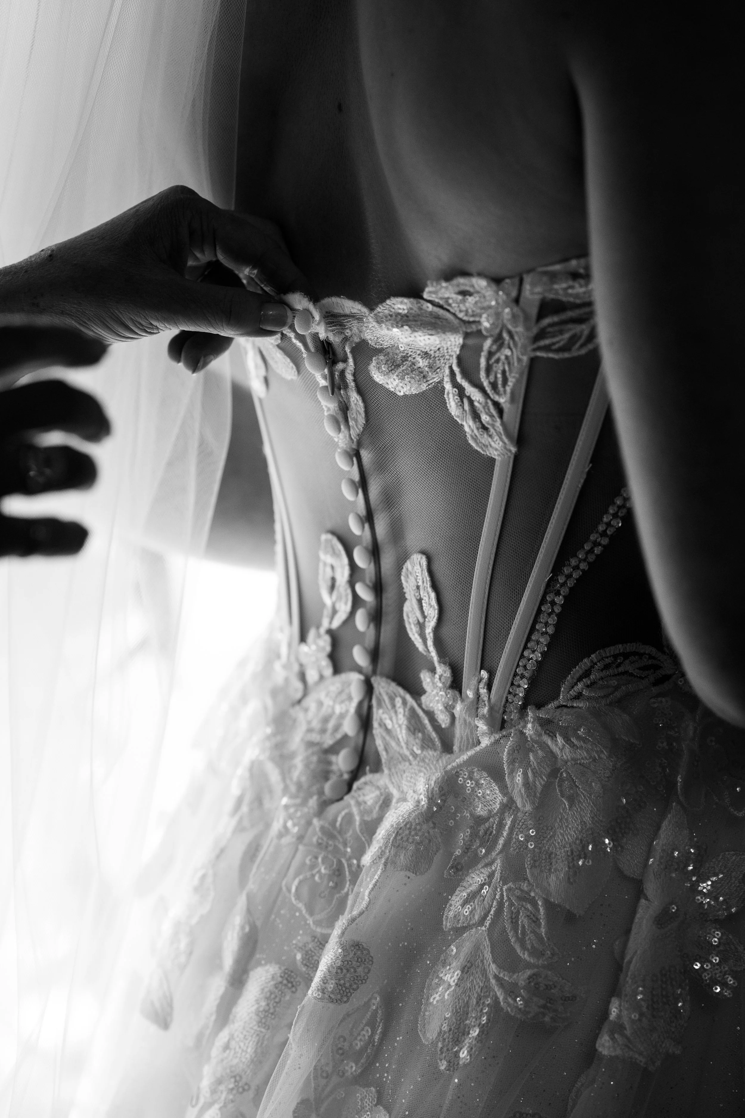 A black and white photo of a bride and groom kissing in a city parking lot, with a large historic building and cars in the background.