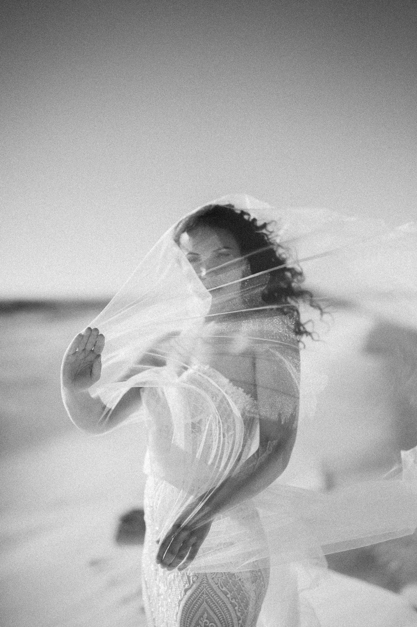 Black and white photo of a woman standing on a beach, wrapped in a veil or shawl, with wind blowing her hair and cloth, against a clear sky.