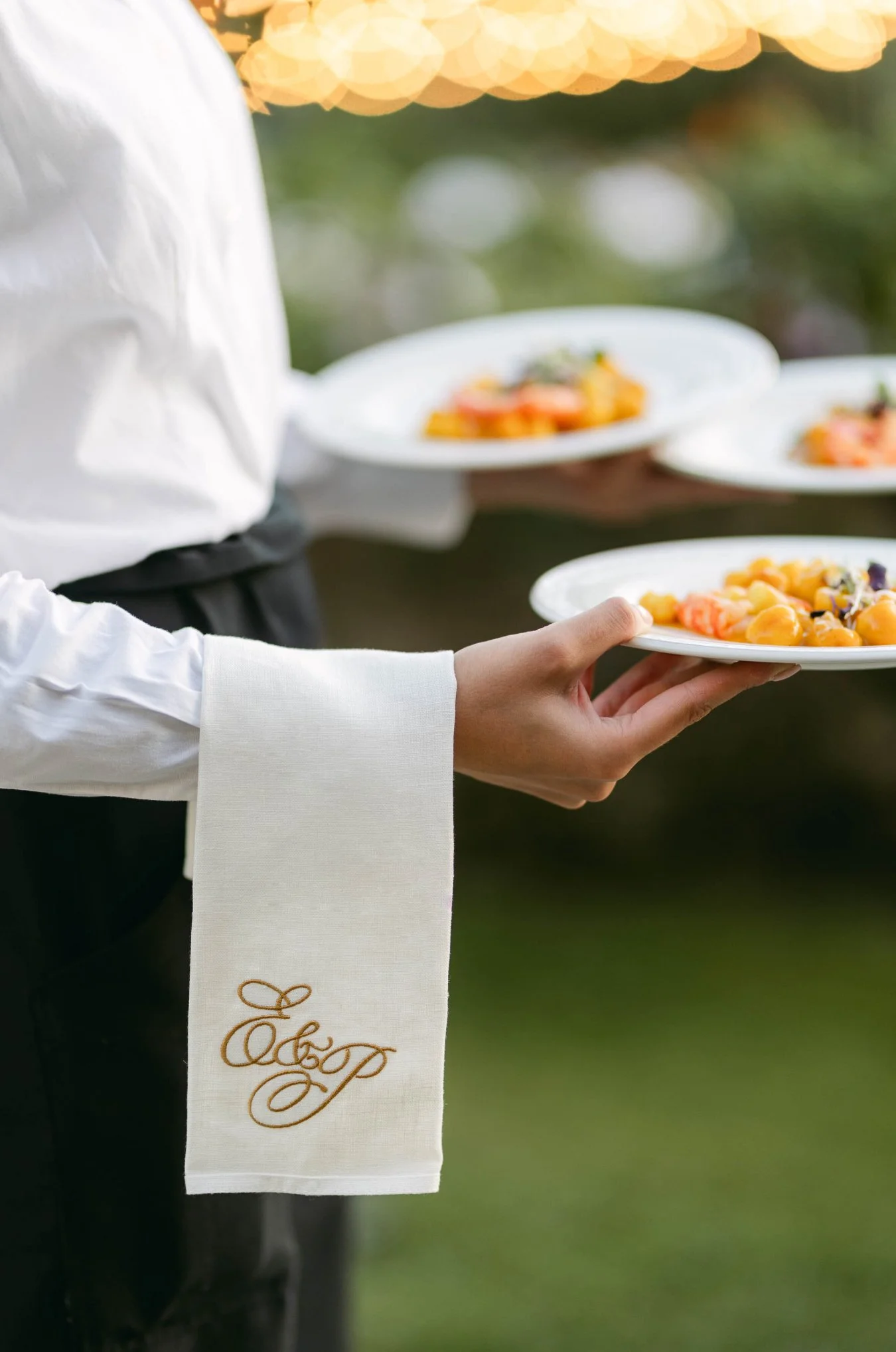 A server dressed in white holds multiple white plates of food outdoors, with a blurred background and bokeh lights overhead.