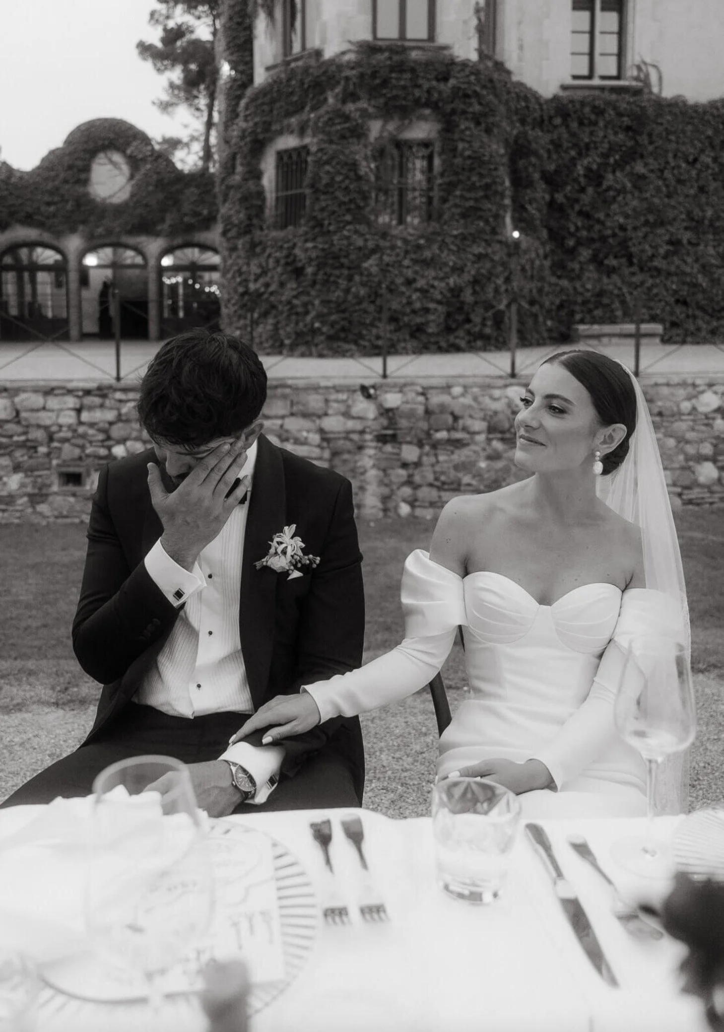 A black-and-white photo of a bride and groom at their wedding, sitting outdoors. The bride, wearing a white off-shoulder wedding dress and veil, gently holds the groom's hand. The groom, in a tuxedo, covers his face with his hand, appearing emotional. There are tables with plates and glasses in the foreground, with a background of a building covered in ivy.