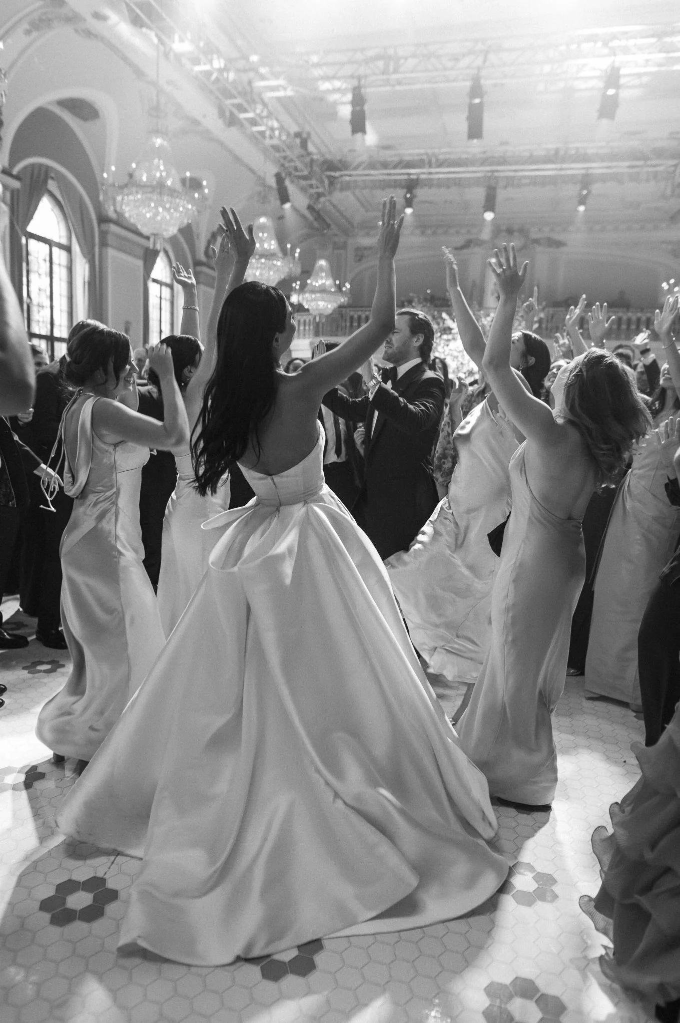 A group of women in elegant dresses dancing with a man in a tuxedo at a formal event in a decorated ballroom.