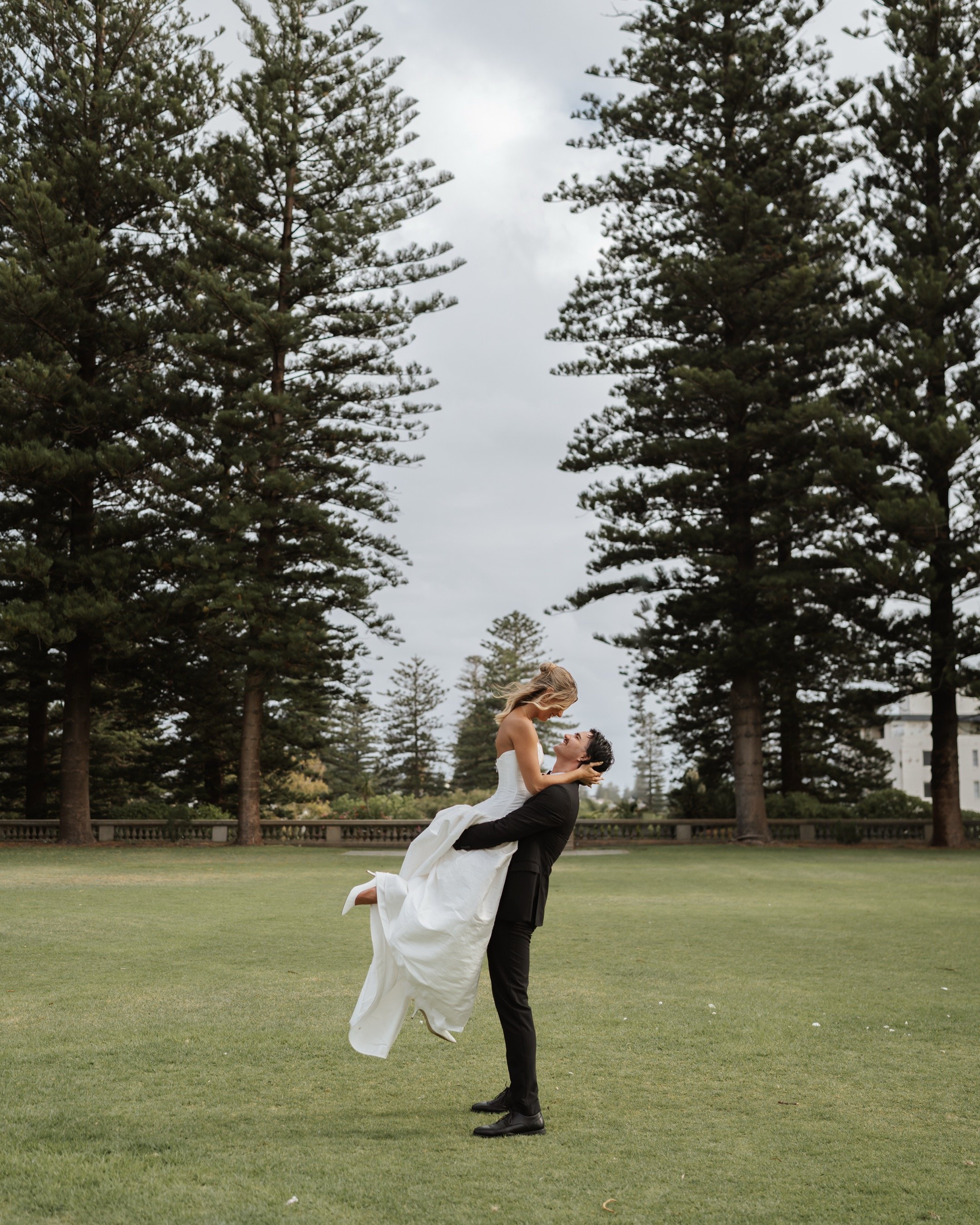 A man in a black suit lifting a woman in a white wedding dress in a green park with tall pine trees.