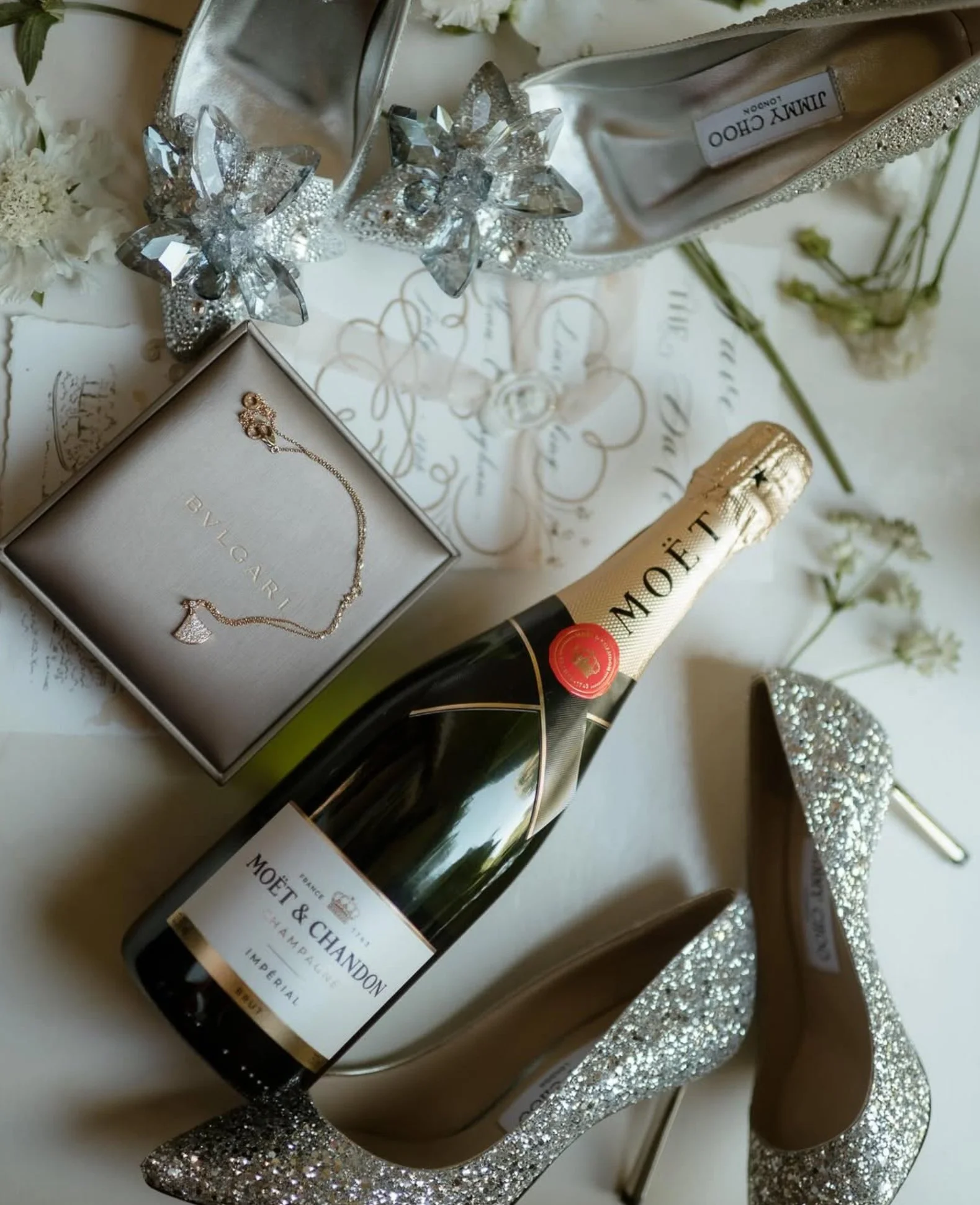 A pair of sparkling high-heeled shoes, a bottle of Moët & Chandon champagne, a jewelry box with a necklace, a decorated shoe with flowers, and floral arrangements on a white surface.
