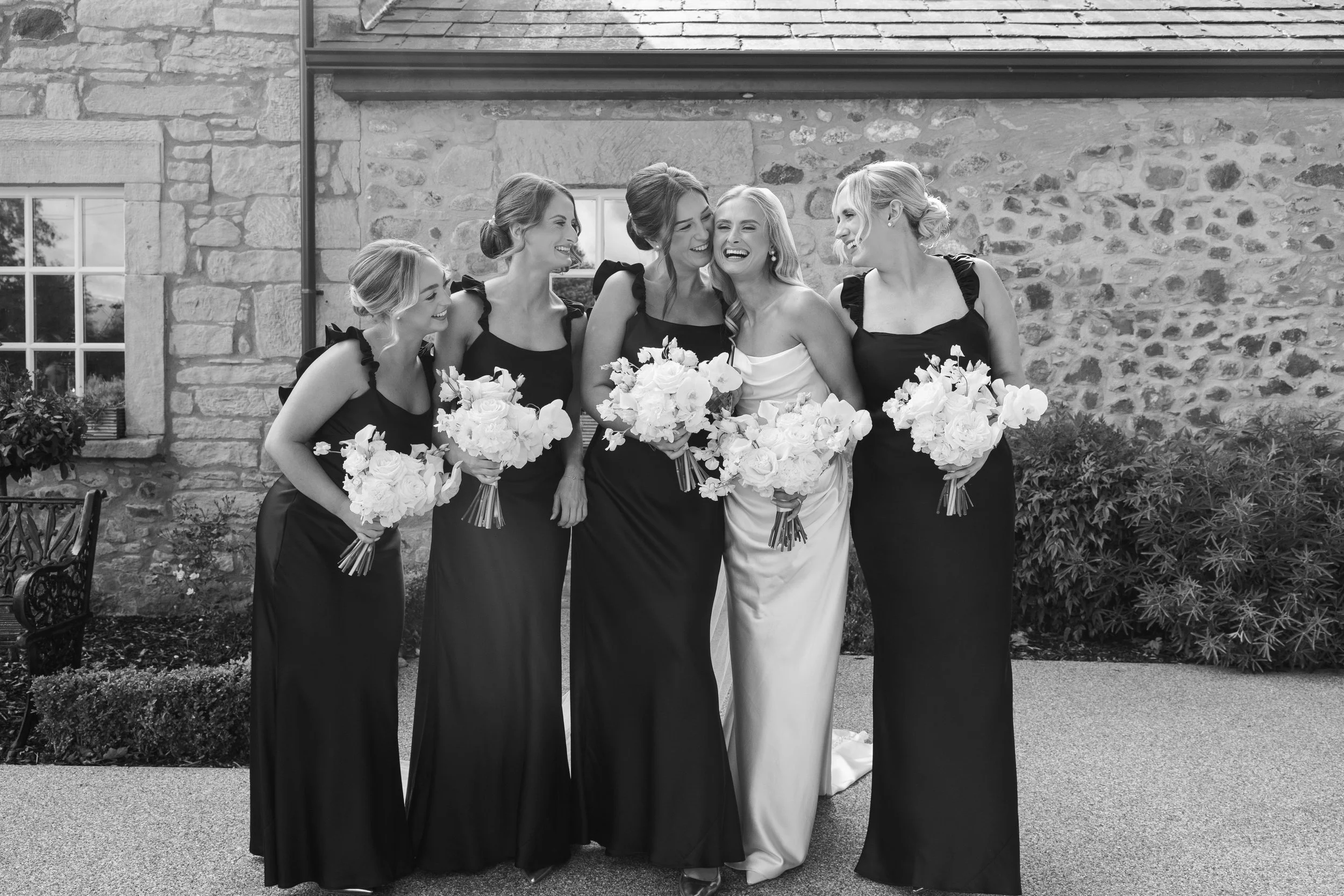 A group of six women, including a bride and bridesmaids, smiling and holding bouquets, standing outside in front of a stone wall with greenery.