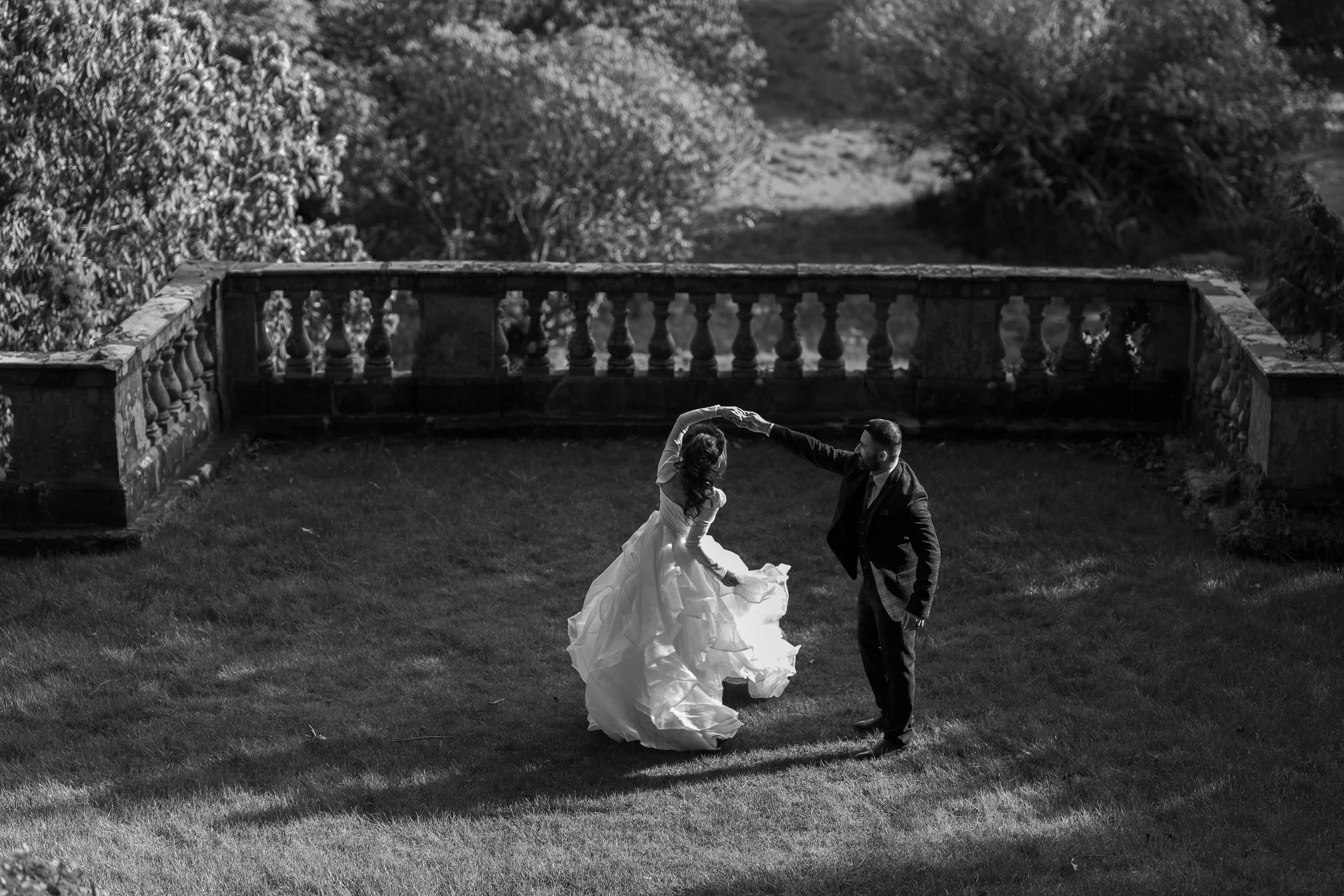 A couple dancing on a grassy lawn, with the woman in a flowing wedding gown and the man in a suit, in front of an ornate stone railing, in a black and white photograph.