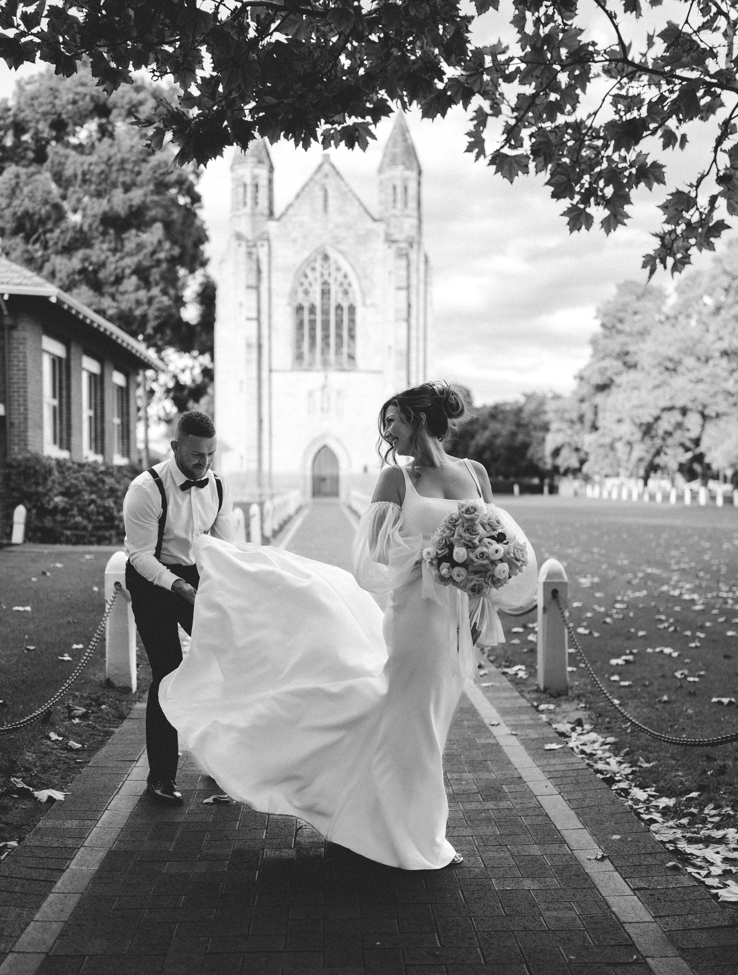 Black and white photo of a bride in a wedding dress holding a bouquet of flowers, smiling and looking to the side, while a man in formal attire carries her dress behind her on a brick pathway outside a church.