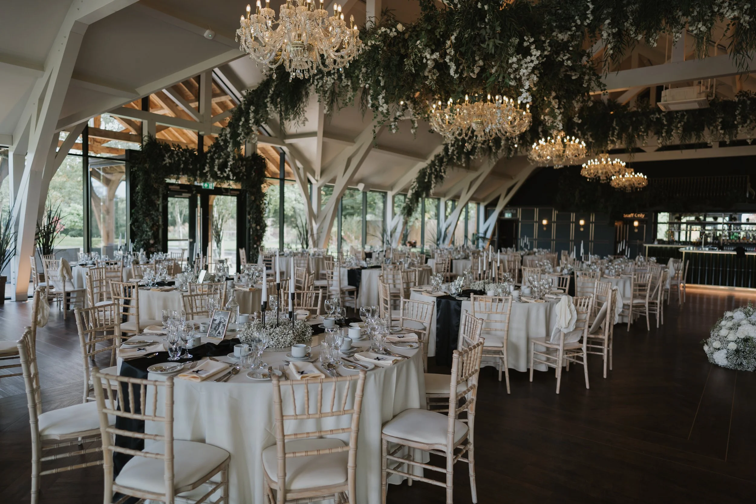 Elegant event space with round tables decorated with white tablecloths, black napkins, floral centerpieces, and glassware. Chandelier lighting, large windows showing greenery outside, and hanging greenery decorations create a luxurious atmosphere.