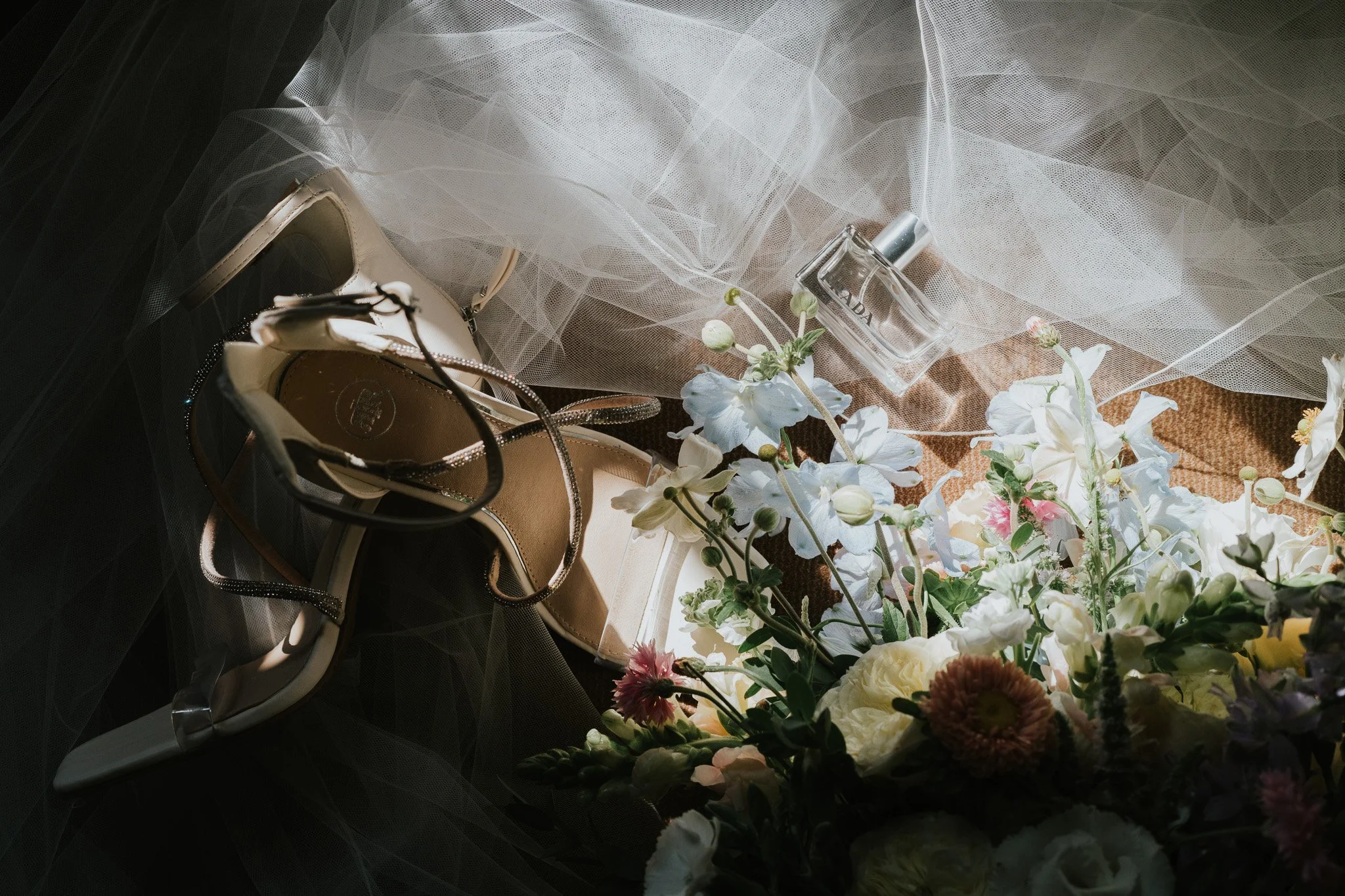 A pair of white high-heeled shoes, a bottle of Prada perfume, a cluster of white and pink flowers, and a piece of white tulle fabric lying on a surface.