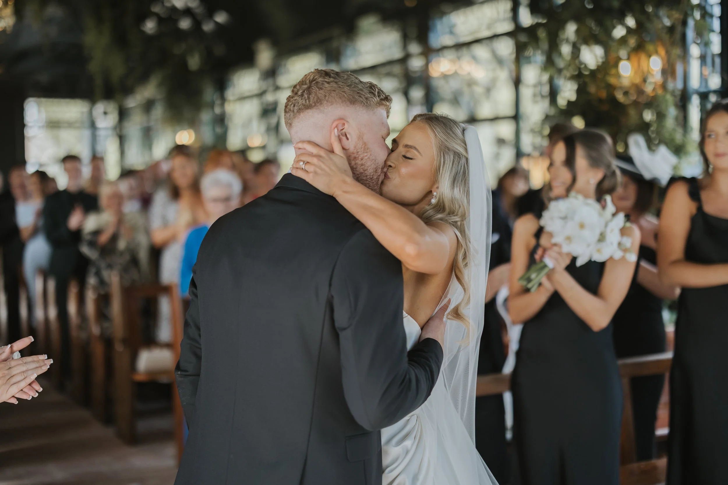 A bride and groom kiss during their wedding ceremony, with guests in the background applauding and holding flowers.