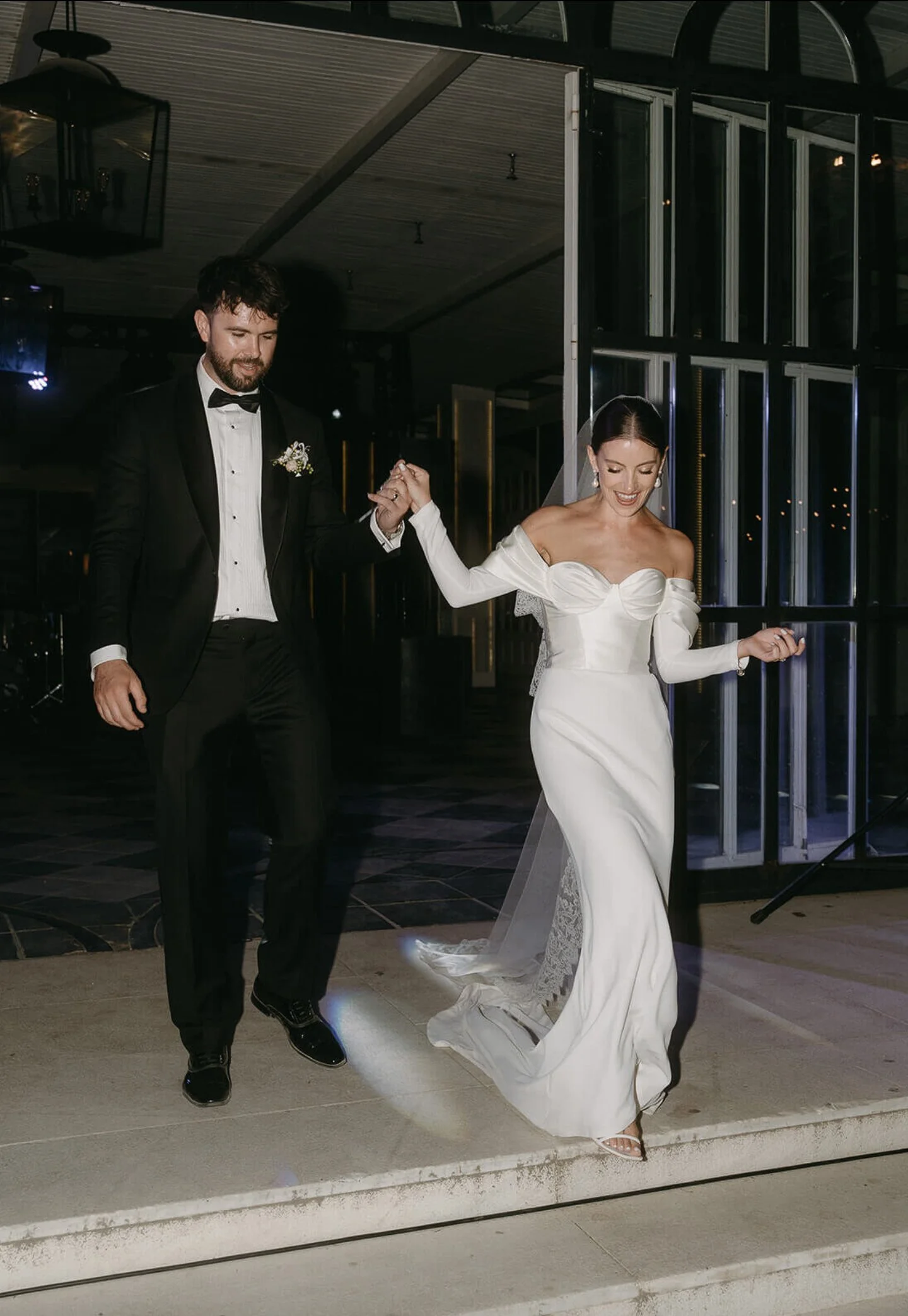A newlywed couple dancing at their wedding reception, with the groom in a black tuxedo and the bride in a white off-the-shoulder wedding gown and veil.