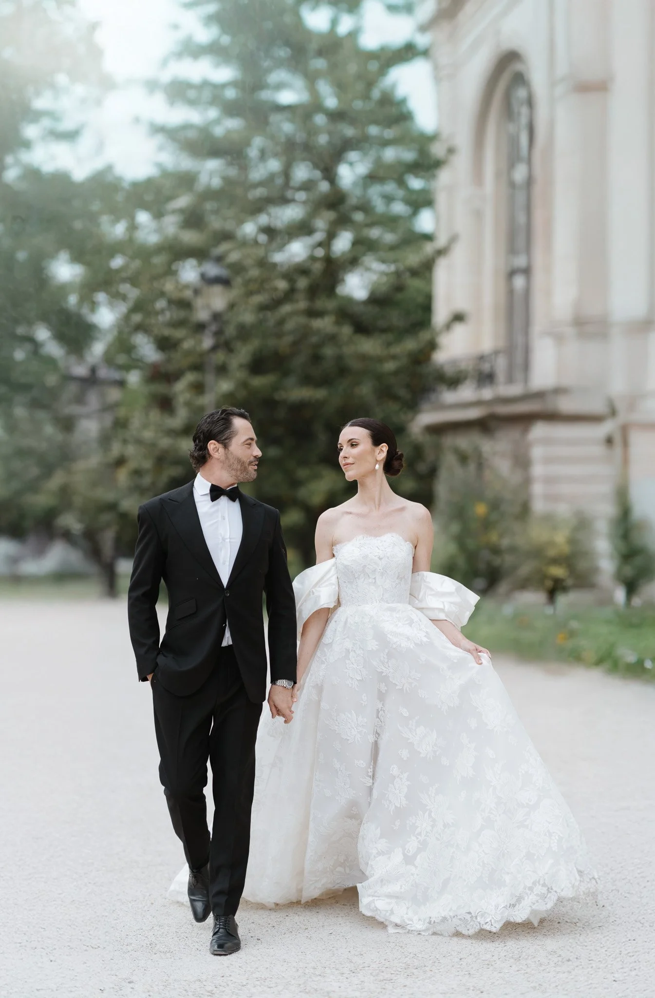 A bride and groom walking hand-in-hand outdoors in front of a historic building, with trees in the background.