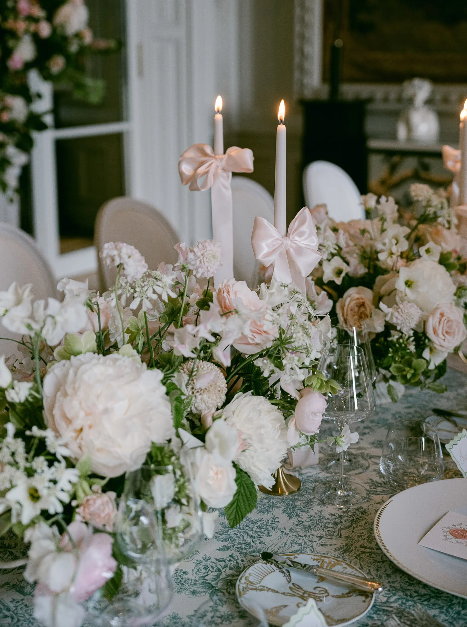 Elegant table setting with a floral centerpiece of peonies, roses, and other soft-colored flowers, lit taper candles with satin bows, white plates with gold trim, and glassware, in a refined indoor setting.