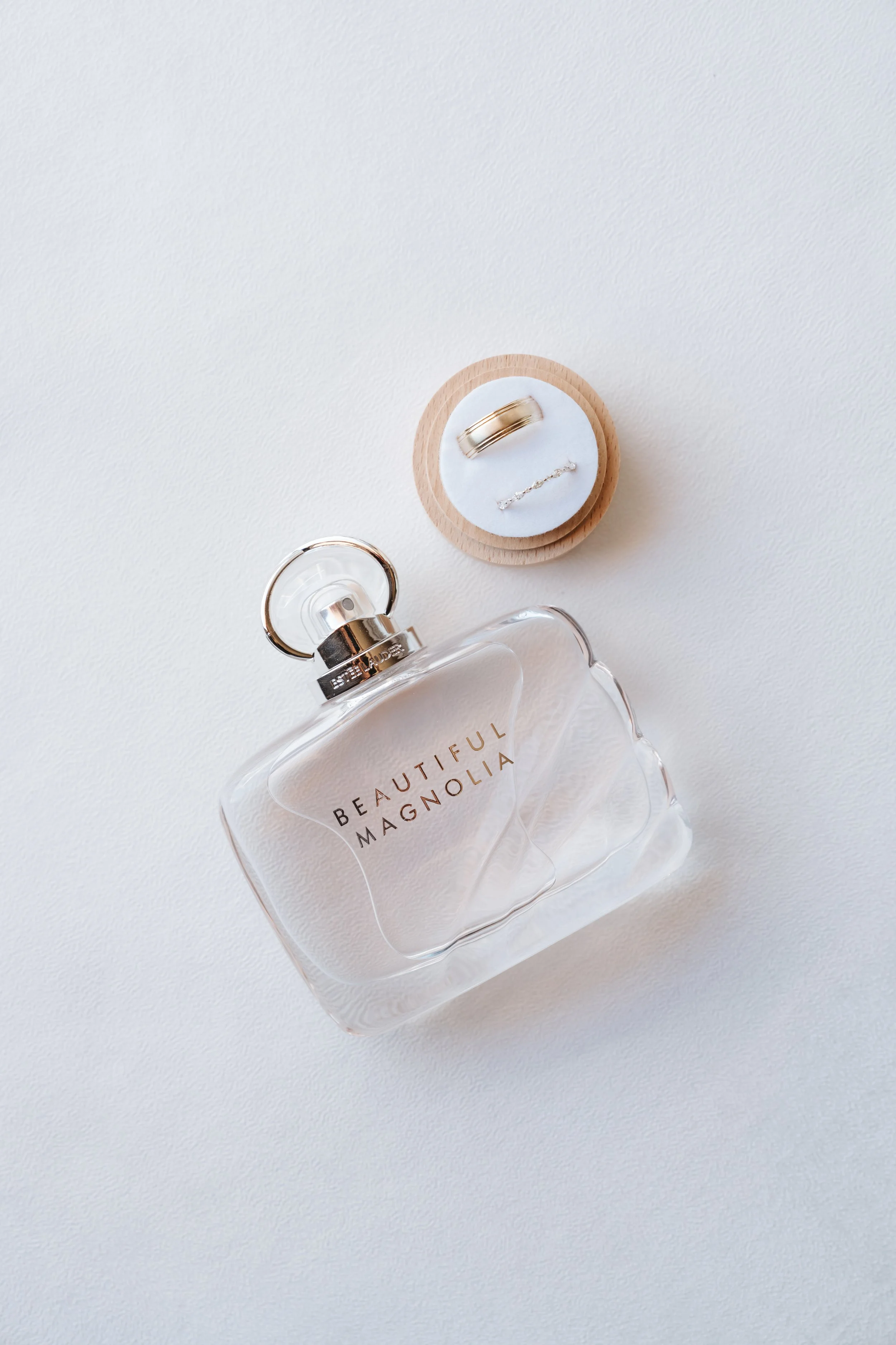 A bottle of perfume labeled 'BEAUTIFUL MAGNOLIA,' a jewelry ring, and a bracelet inside a small round wooden dish on a white surface.