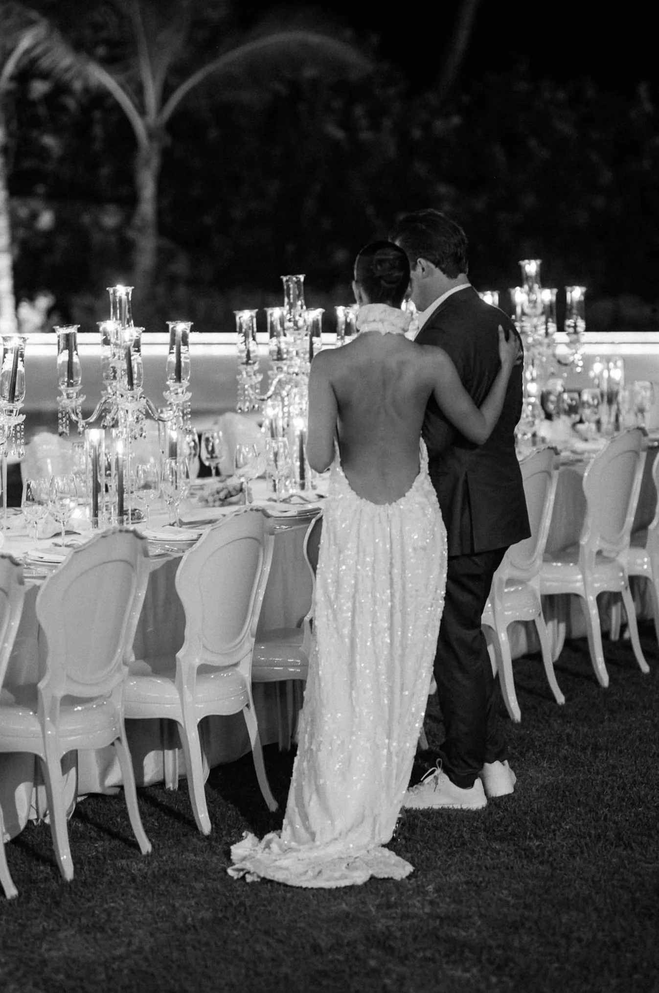 A black and white photo of a bride and groom dancing at a wedding reception outdoors at night, surrounded by a long table set with candles and glassware.