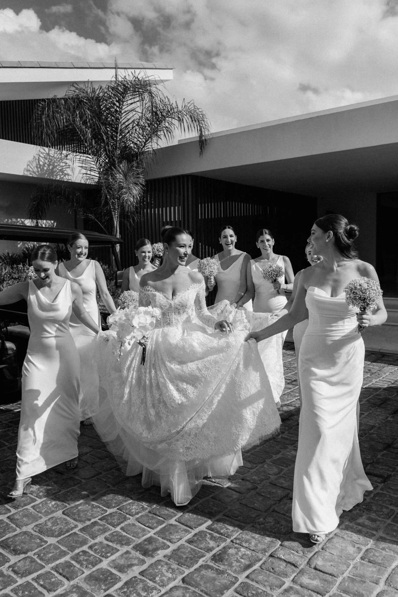 Group of women in wedding dresses and bridesmaids' dresses smiling and walking outdoors on brick pavement, with modern building and palm trees in background.