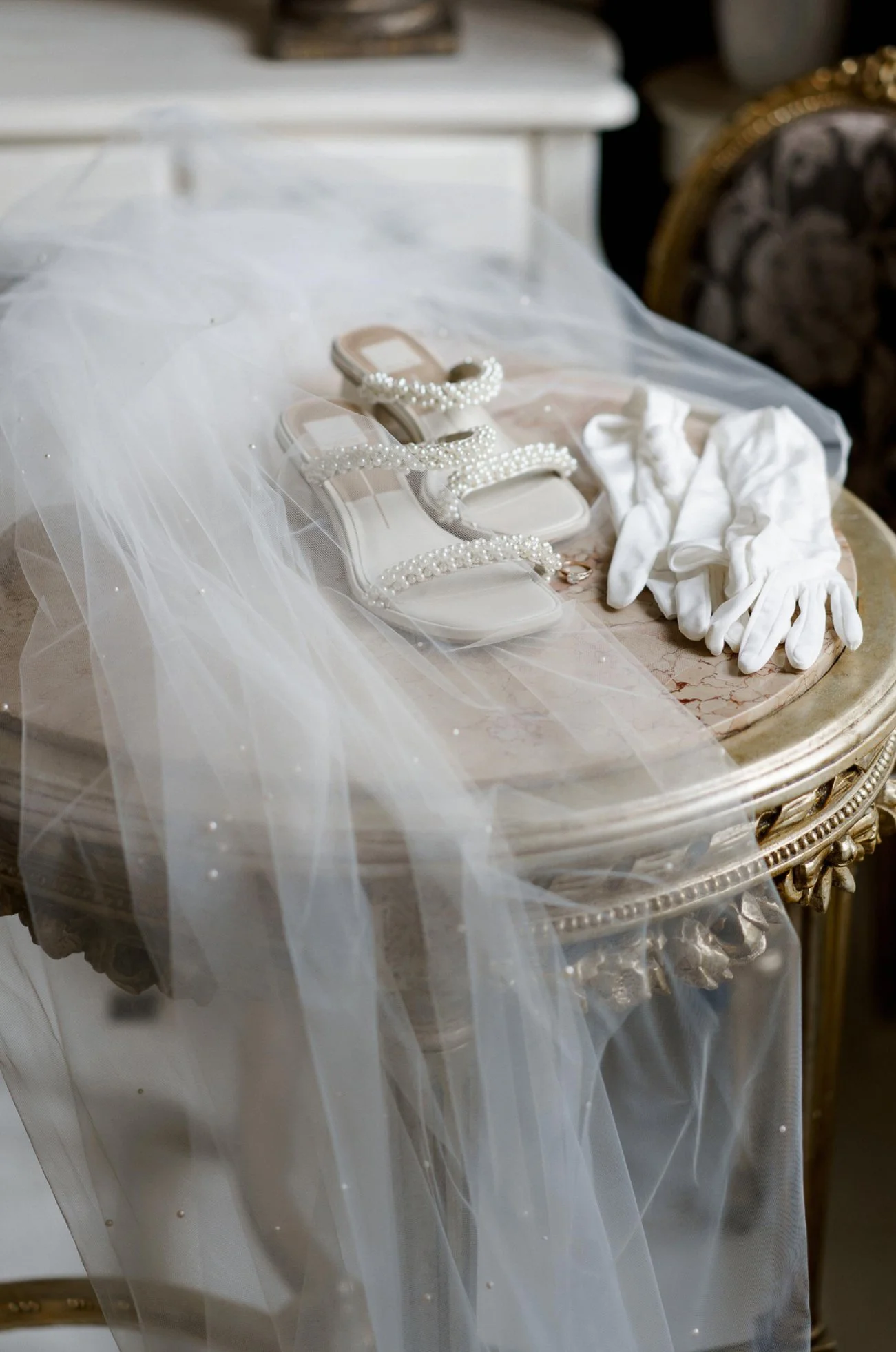 A pair of white wedding shoes with pearl embellishments, white gloves, and a white veil on a vintage ornate table.