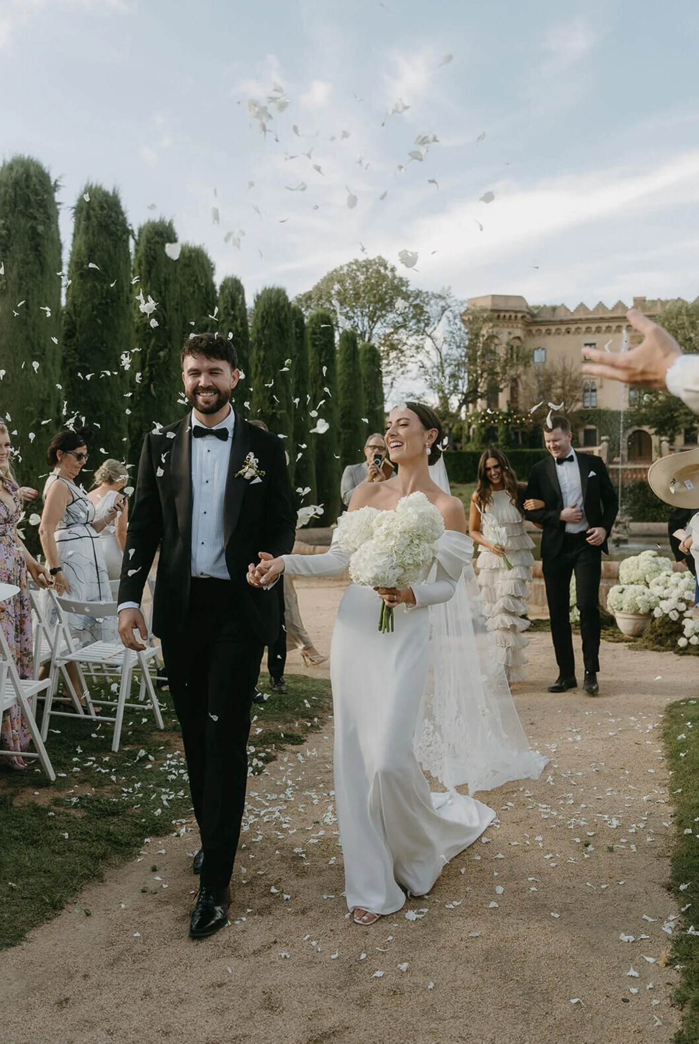 Bride and groom smiling and holding hands at their outdoor wedding ceremony, surrounded by guests, confetti, and floral decorations.