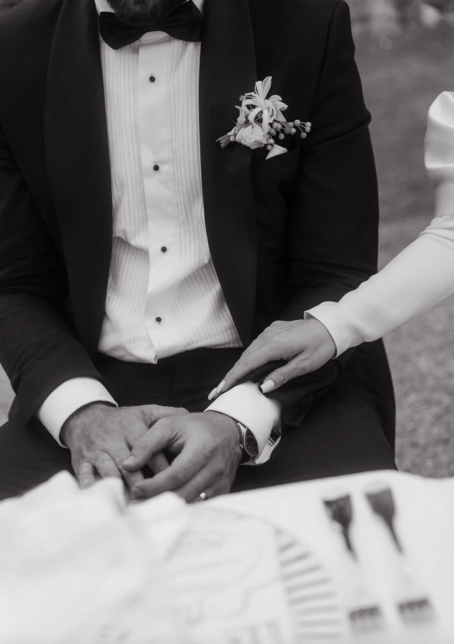 Close-up of a groom in a tuxedo and bow tie, with a floral boutonniere, holding hands with a bride in a light-colored sleeve, touching his arm, at a wedding reception or ceremony.