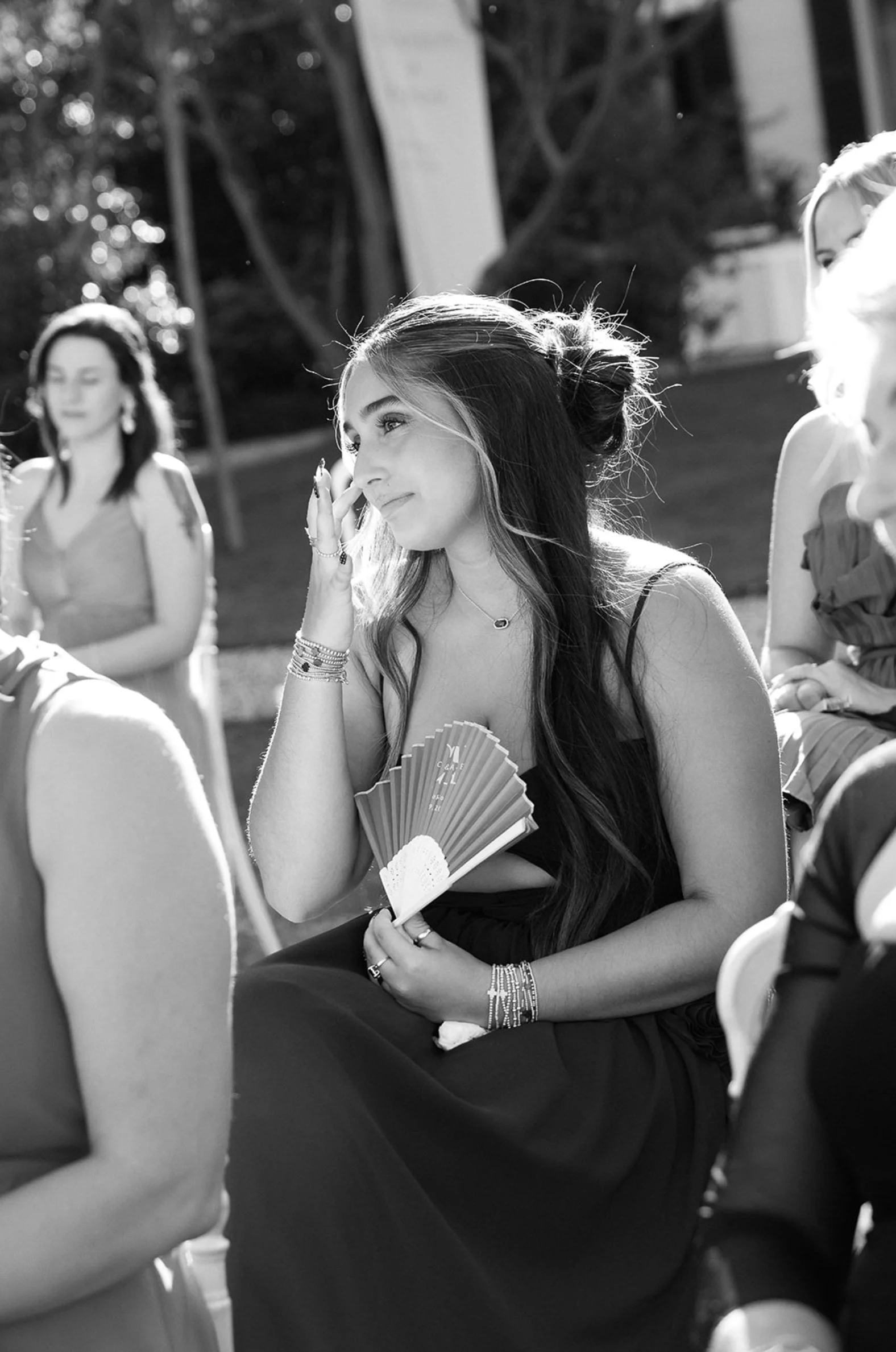 A woman with long hair tied in a bun, sitting outdoors, appears to be wiping a tear from her eye while holding a fan, surrounded by other women at a social event.