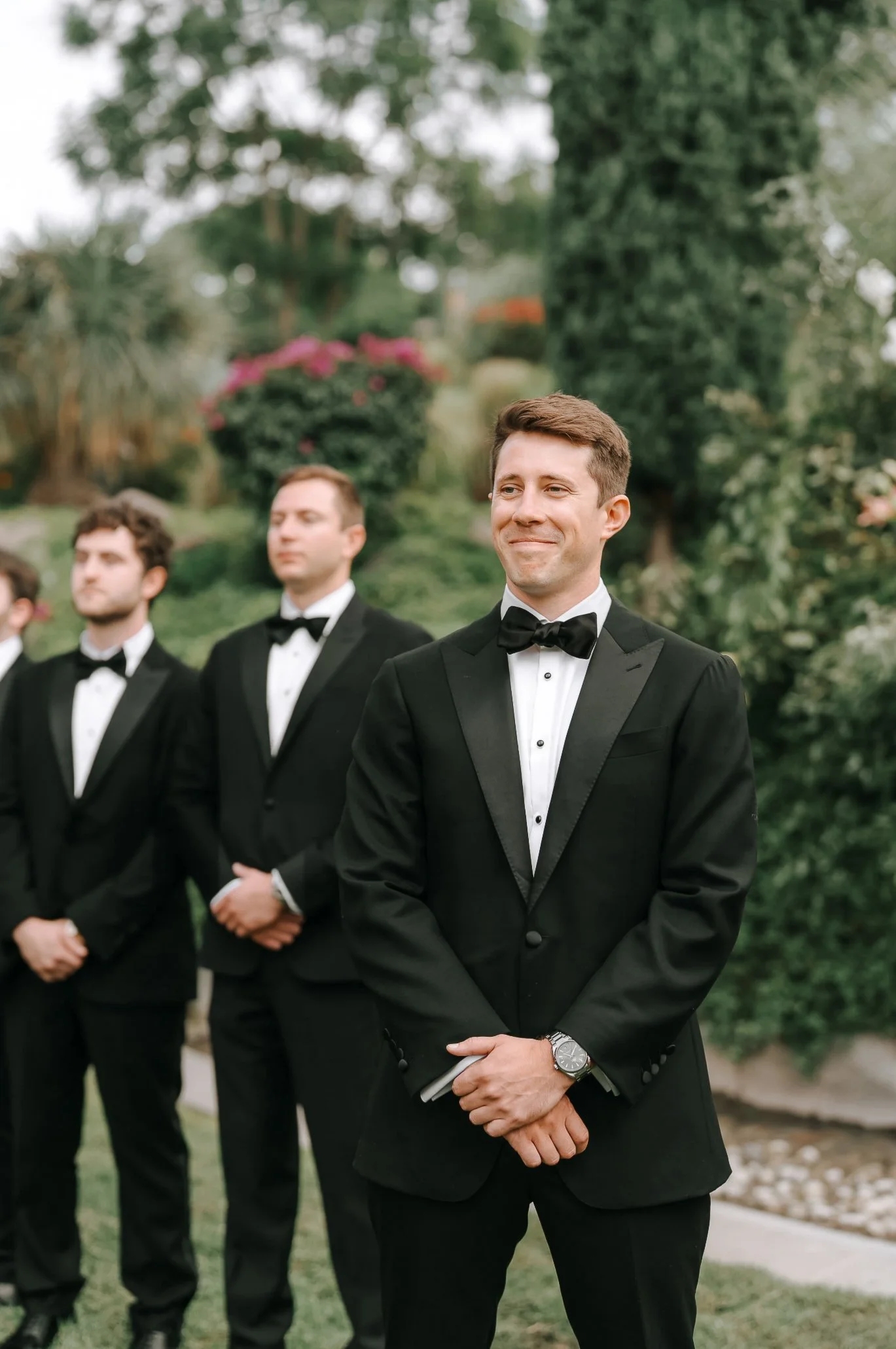 A groom in a black tuxedo and bow tie standing outdoors during a wedding ceremony, smiling with three groomsmen in the background, all dressed in similar tuxedos, amid greenery and trees.
