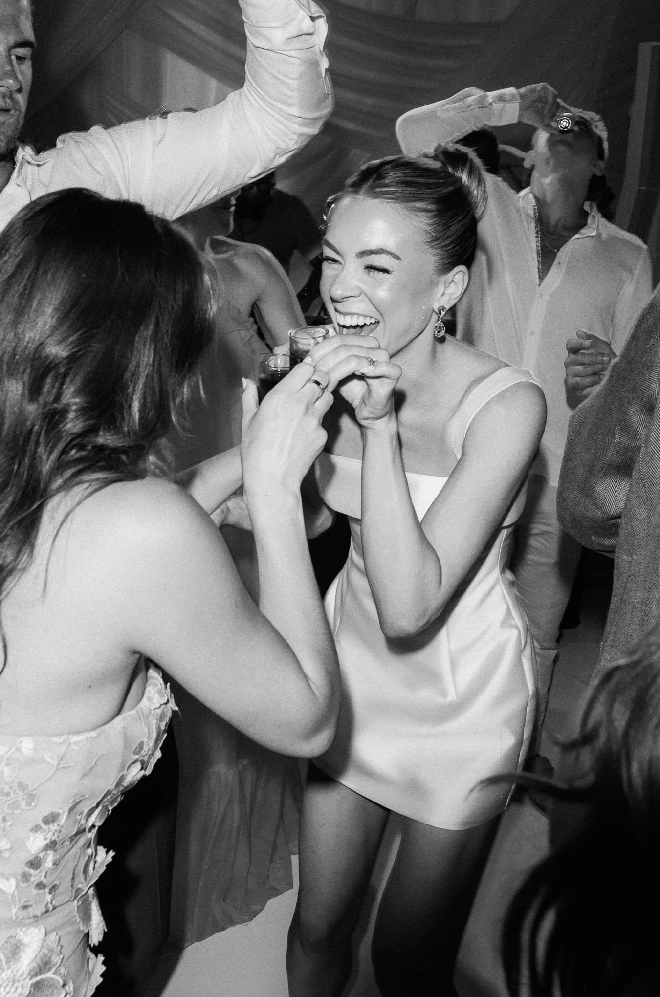 People at a celebration, with a woman in a satin dress laughing and holding a glass, surrounded by others in party attire.