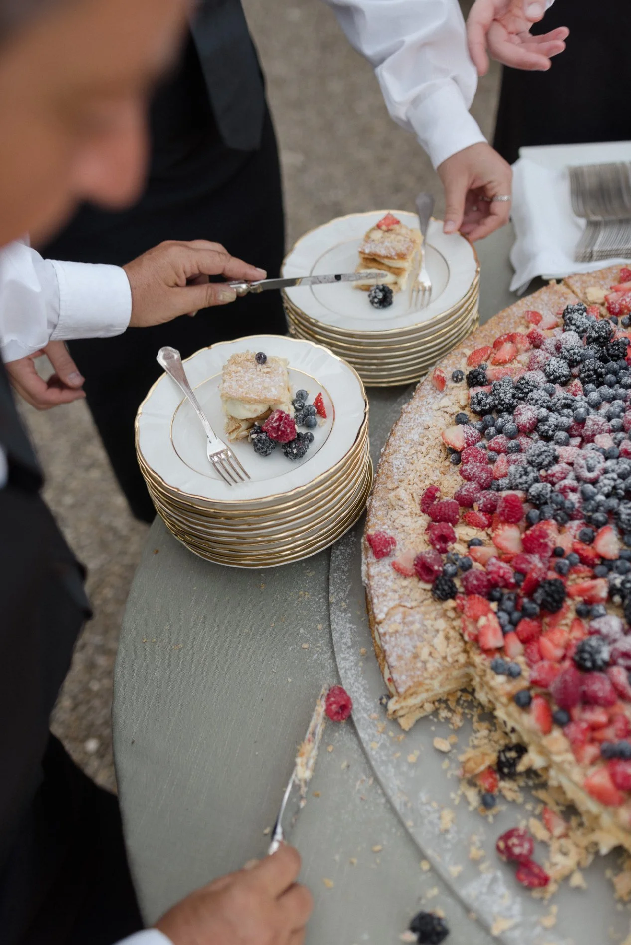 People serving slices of fruit pie onto small plates with berries, next to a large berry-topped dessert.