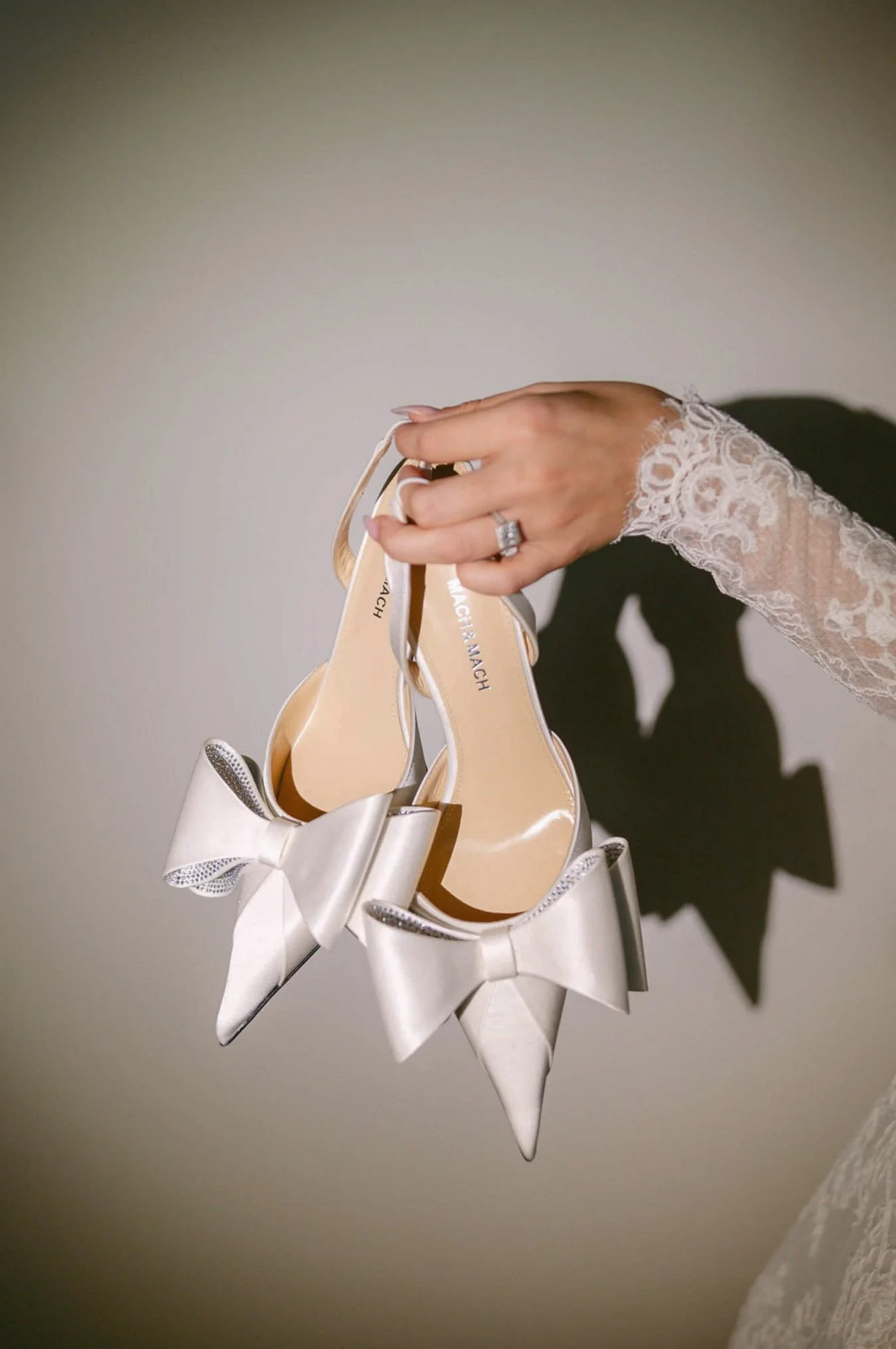 Hand holding a pair of white wedding shoes with bows, lace sleeve, and a diamond ring.